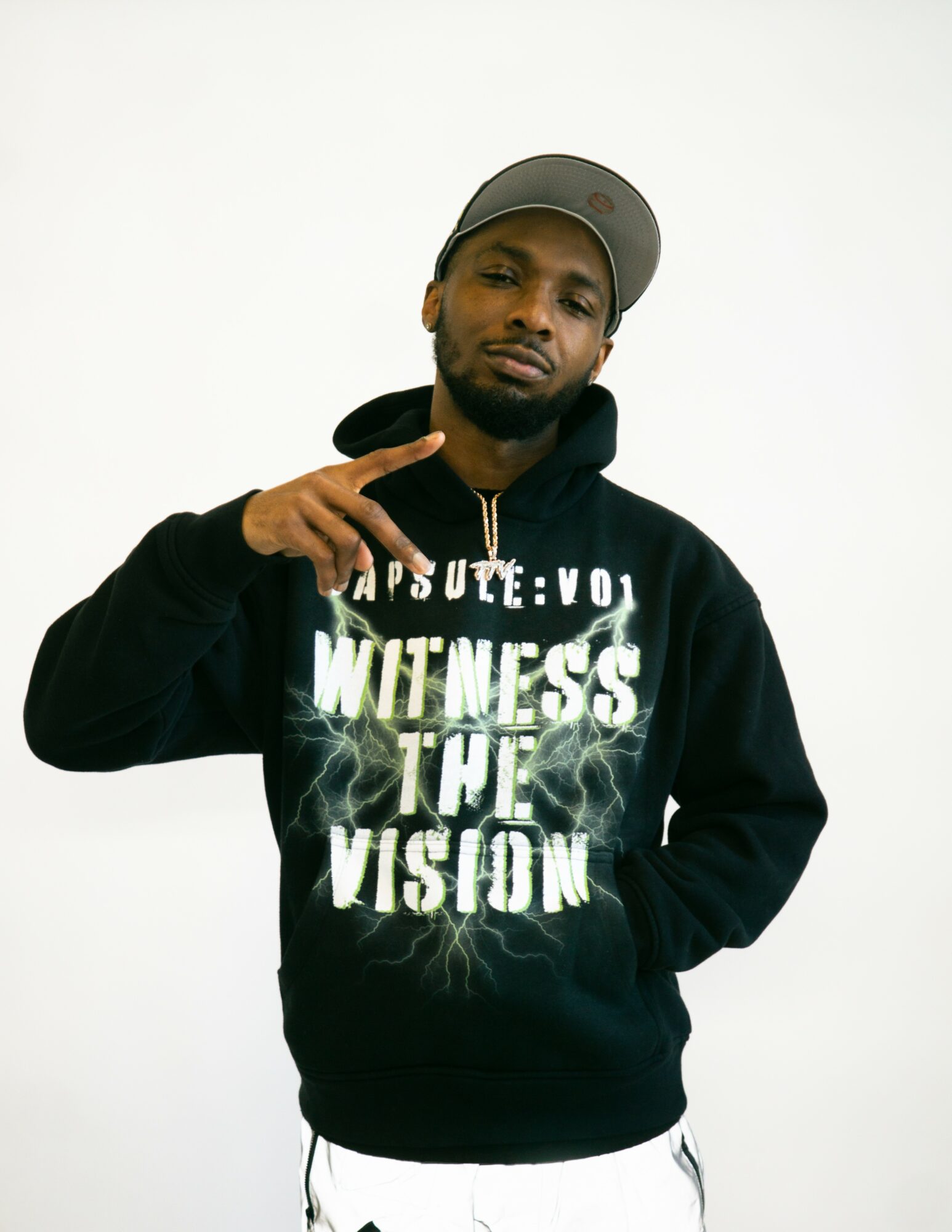 Man wearing a black hoodie and gray cap, making a peace sign with his right hand, against a white background.