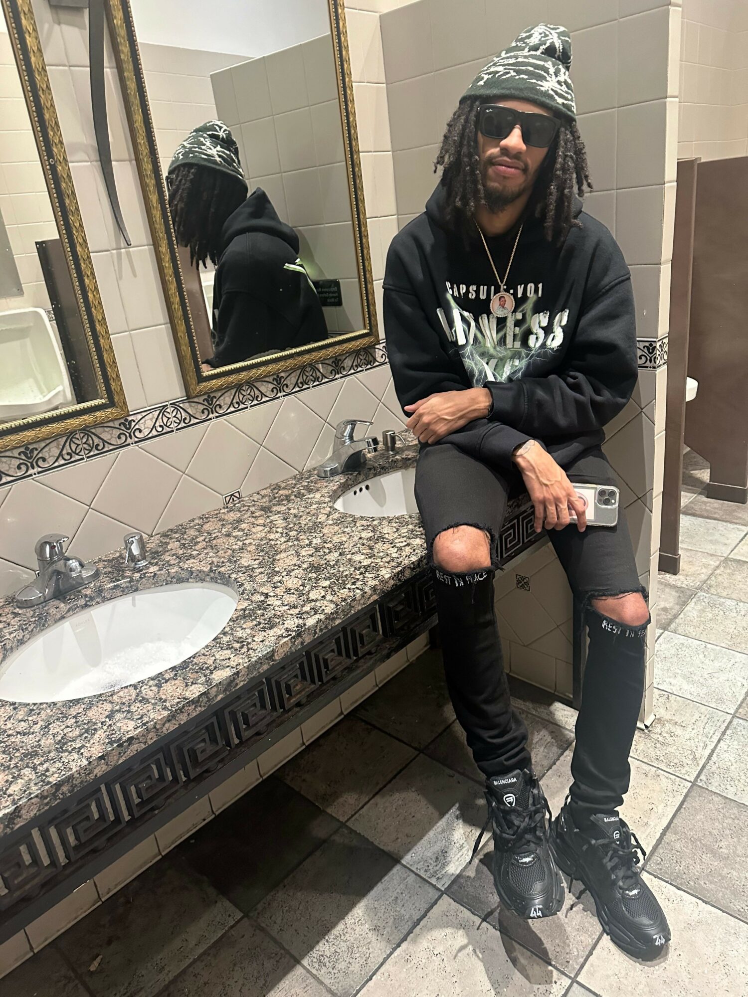 Person taking a mirror selfie in a bathroom, sitting on a countertop with a sink, wearing black clothing and sunglasses.