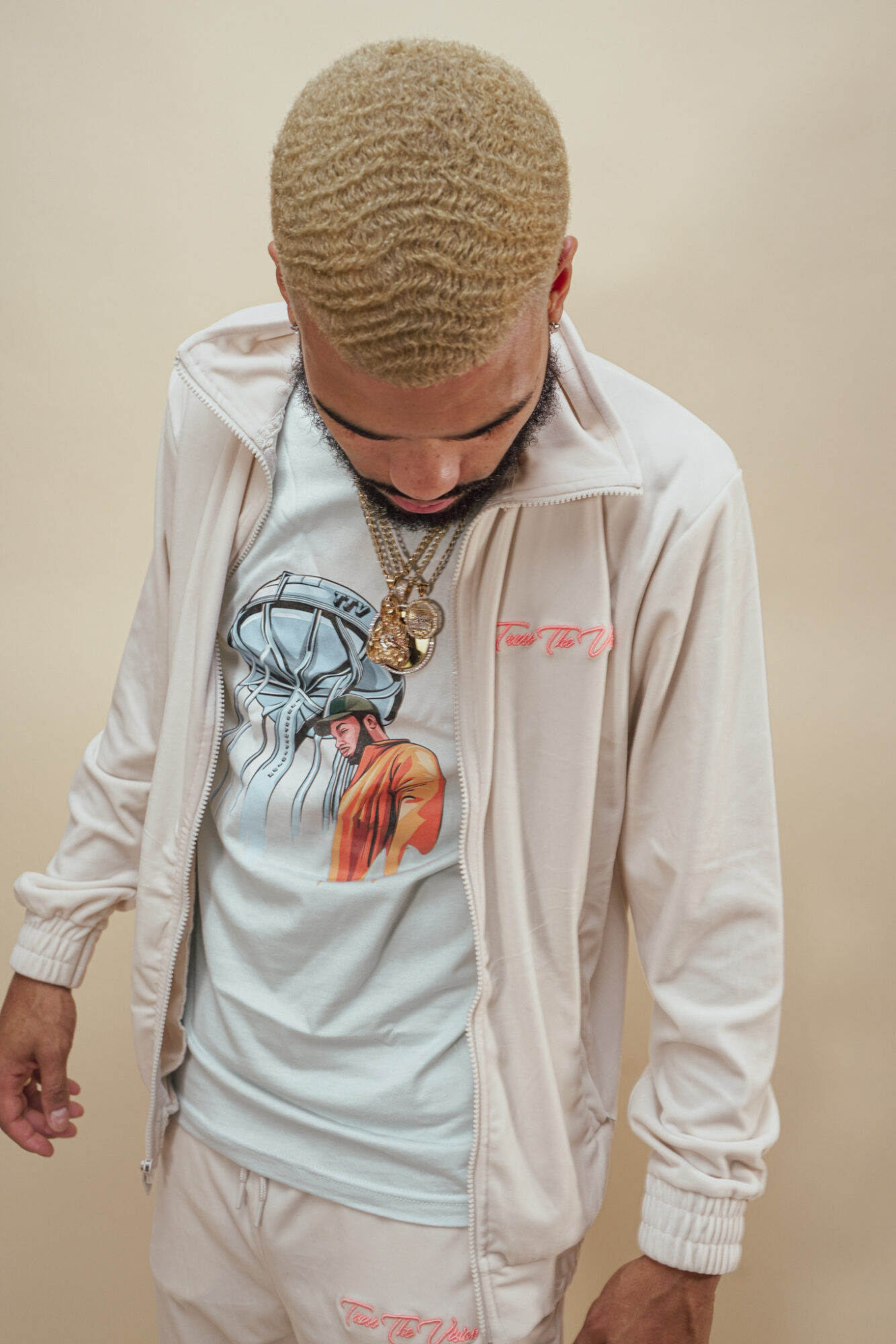 Young person with short, curly blonde hair looking down, wearing a white jacket and graphic t-shirt, against a plain background.