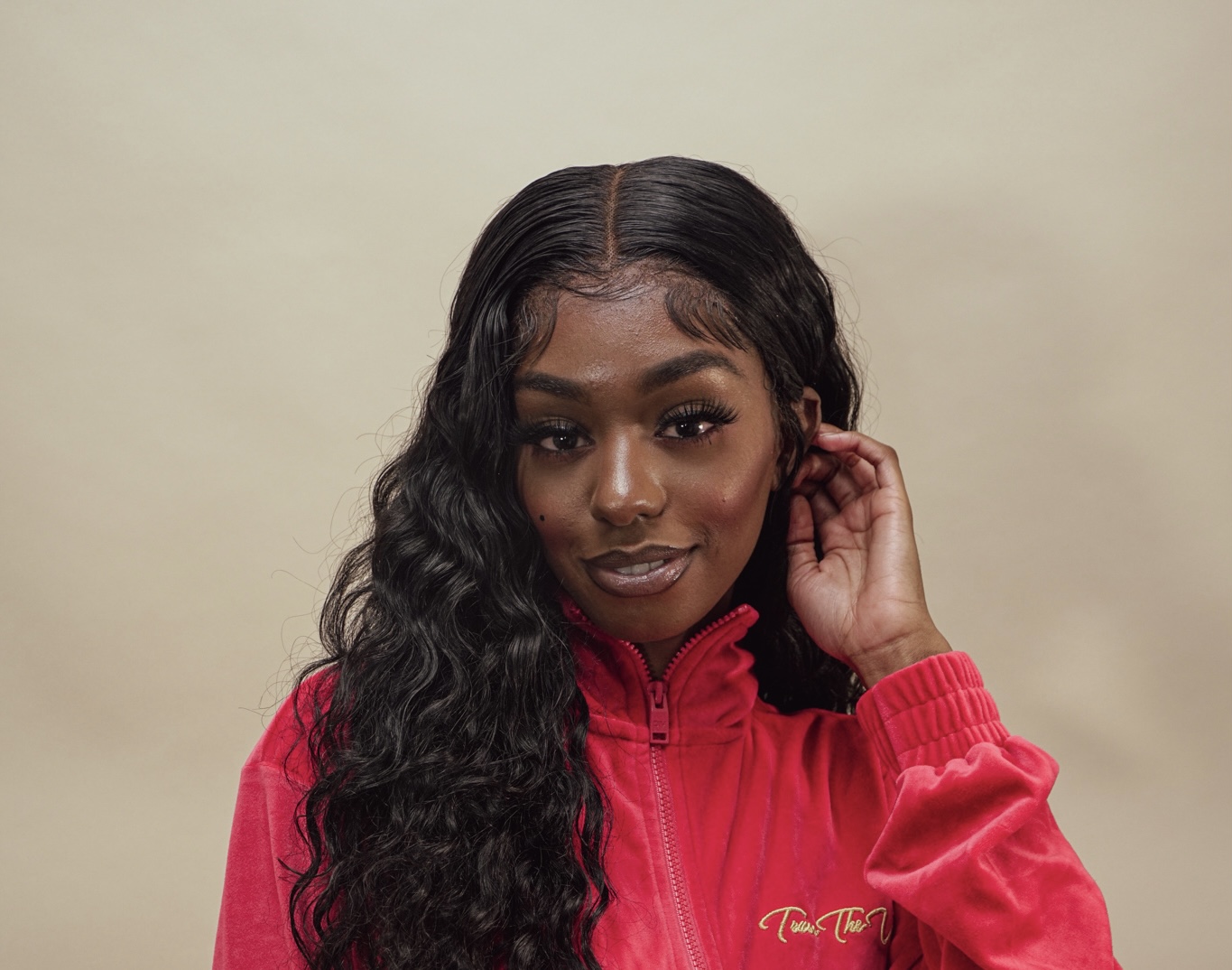 Young woman with long, wavy black hair wearing a red zip-up jacket, touching her ear, smiling softly.