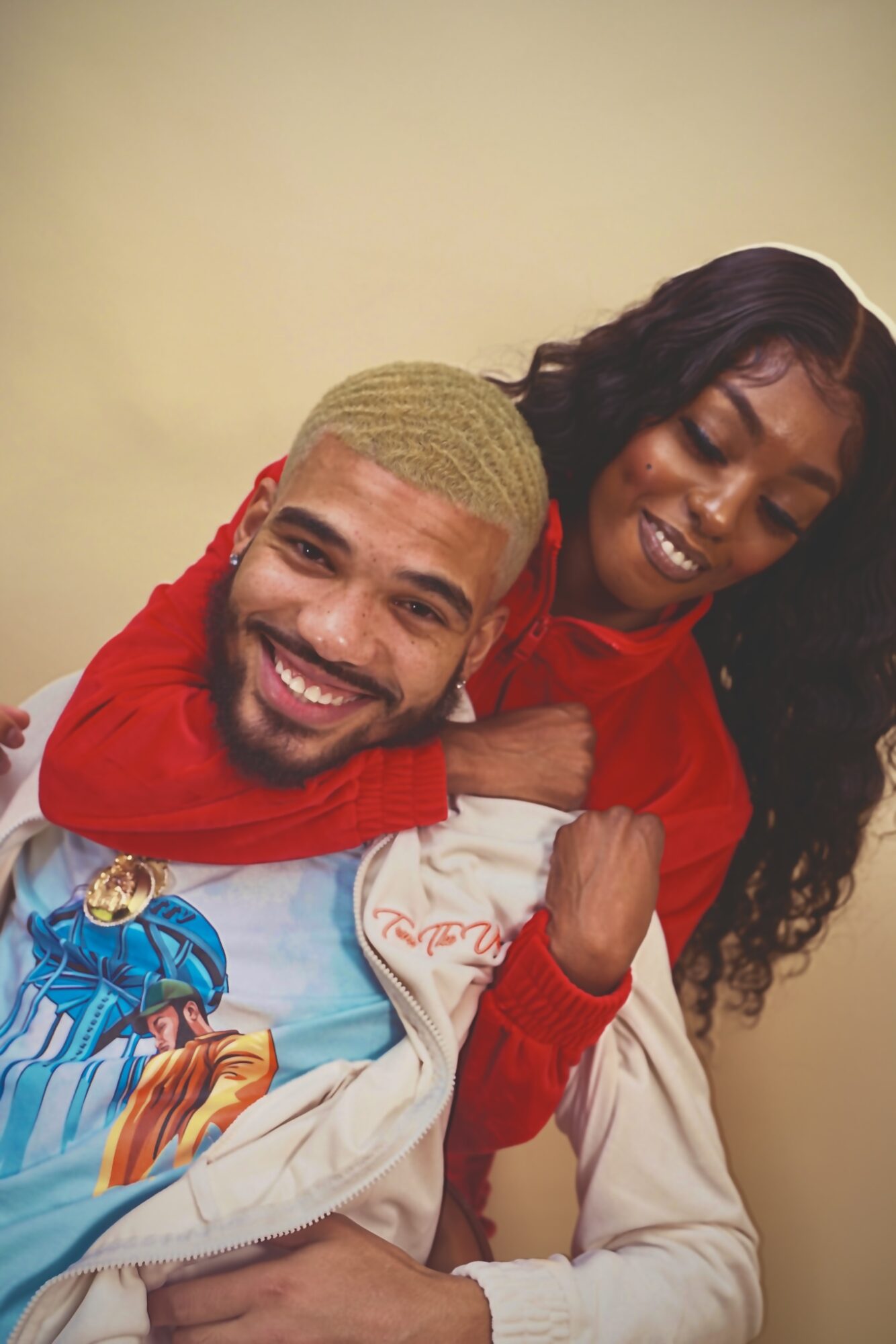 Two people smiling, one hugging the other from behind, both wearing colorful clothing, against a plain background.