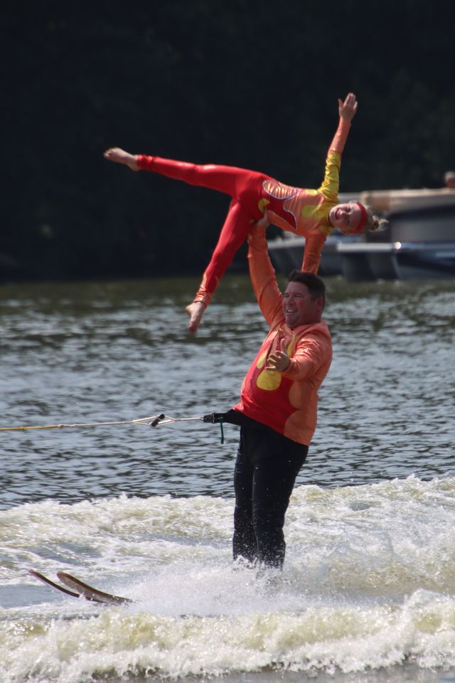 A person in a red shirt holds a performer in a red and yellow costume above their head near water.