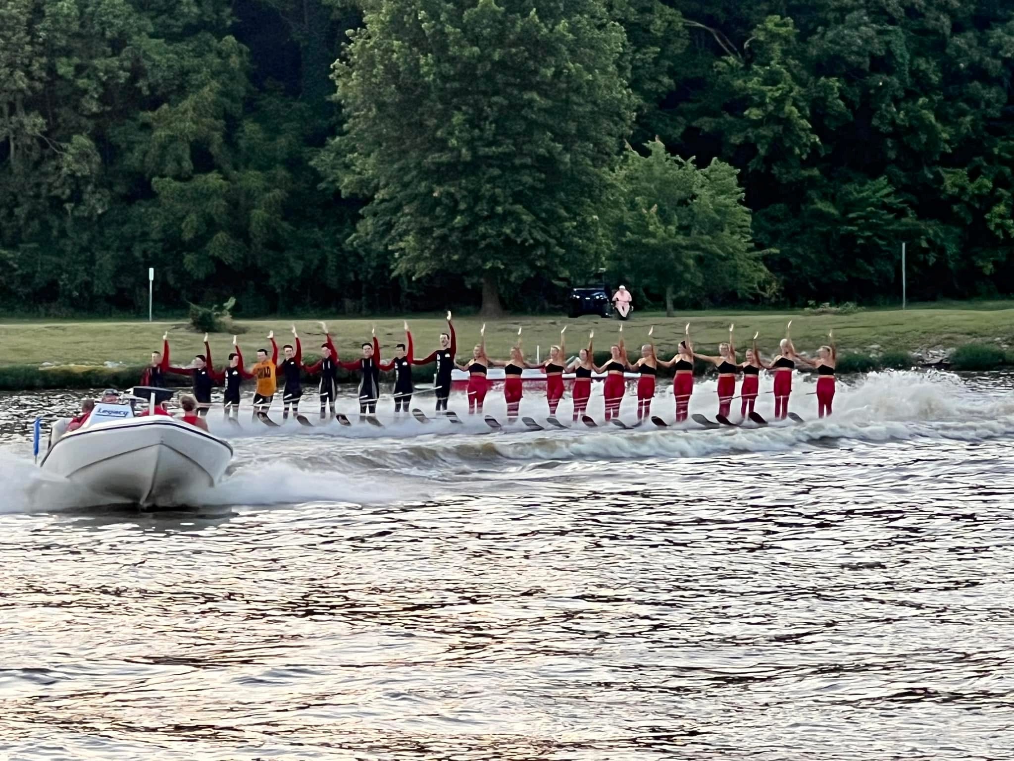 Group of people dressed as Santa Claus and reindeer standing on a boat in water, with trees in the background.