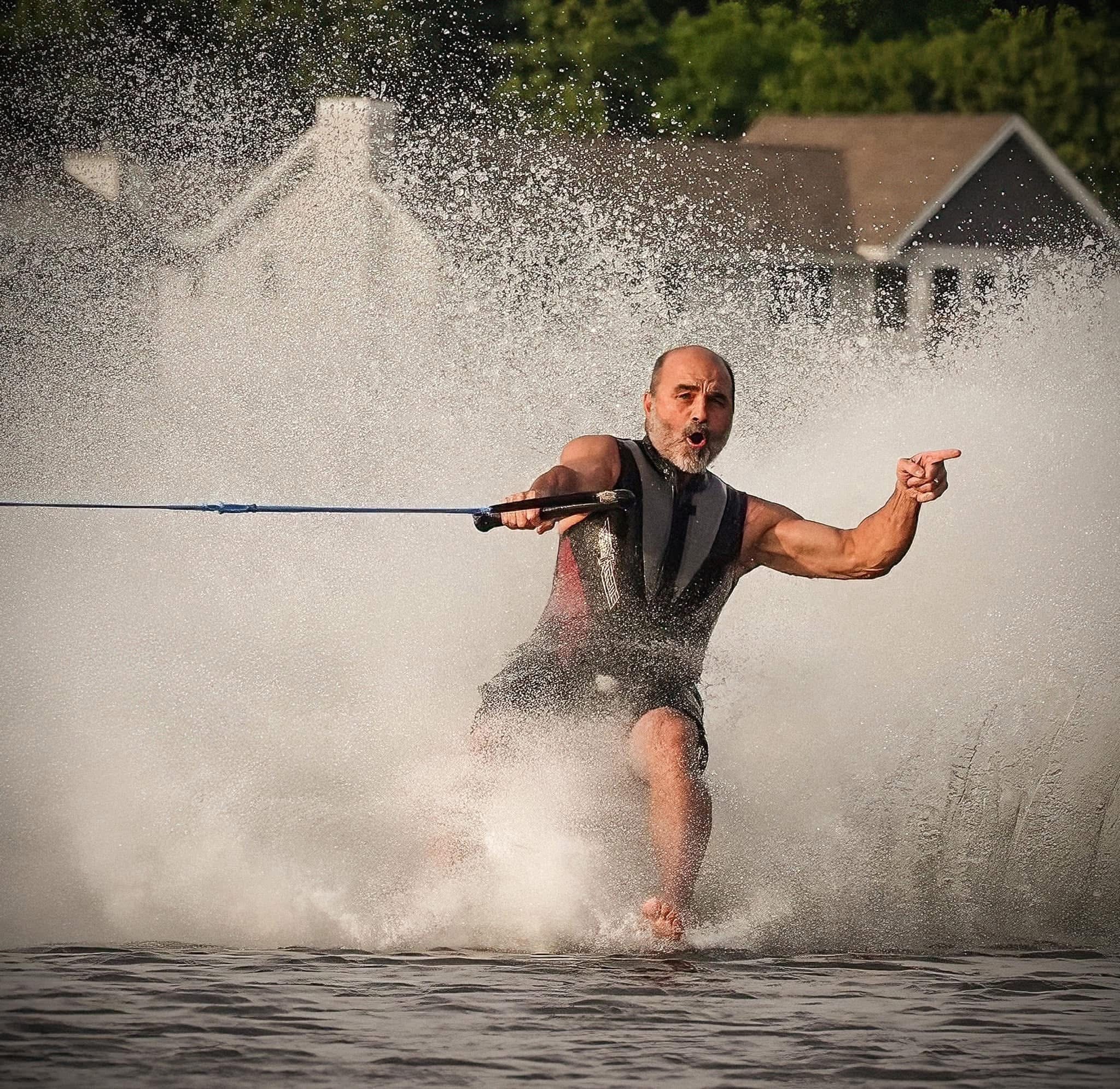 Man wakeboarding on water, holding a rope, with water spray around him, houses and trees in background.