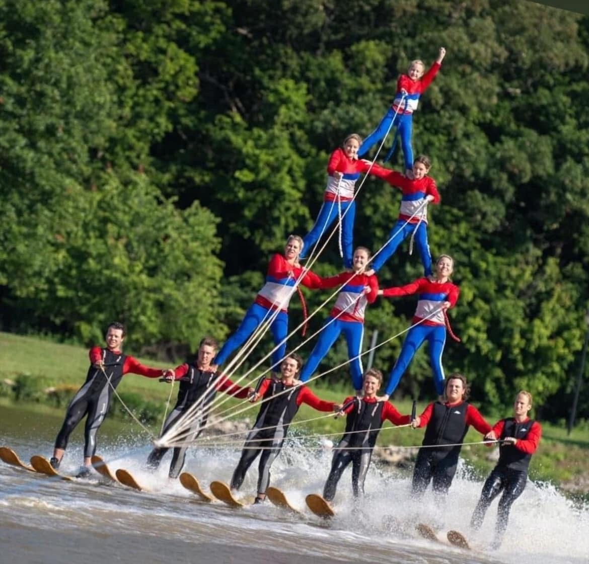 Group of people in life jackets performing a coordinated water-based stunt, forming a human pyramid on skis, with trees in background.
