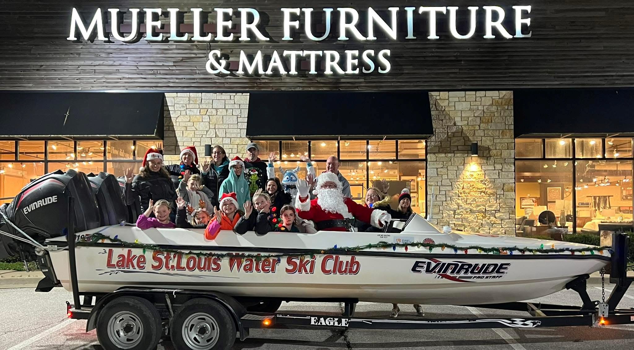 Group of people, some dressed as Santa, on a boat in front of a store with a sign reading 'Mueller Furniture & Mattress'.