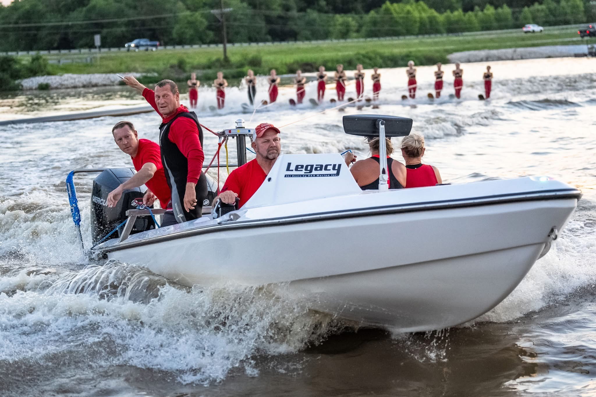 Four people on a white motorboat speeding on water, with a line of people in red shirts in the background.