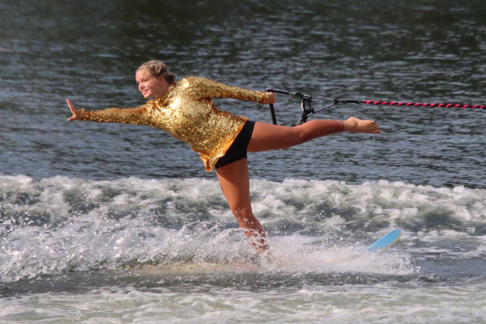 Girl in gold jacket and black shorts balancing on a surfboard in water, holding a rope behind her, with waves around.