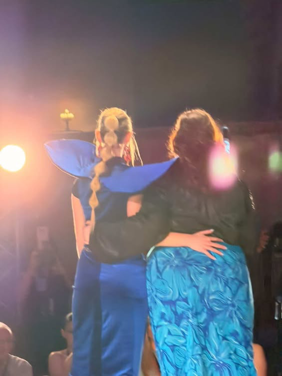 Two people with their backs to the camera, one in a blue costume with a braid, the other in a patterned blue skirt, embracing.