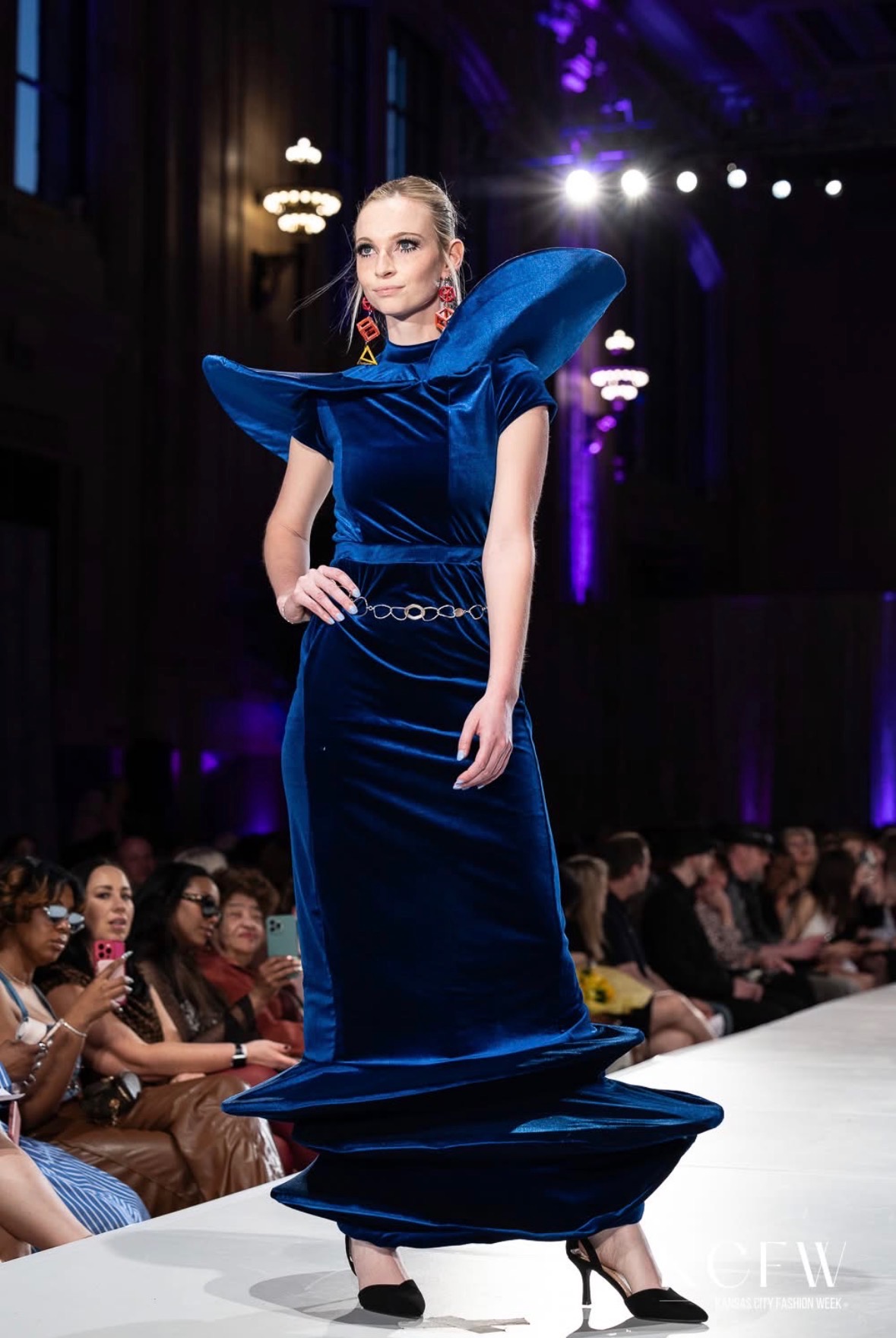 Model walking on runway in a dark blue gown with large shoulder accents, audience seated on both sides, stage lights overhead.