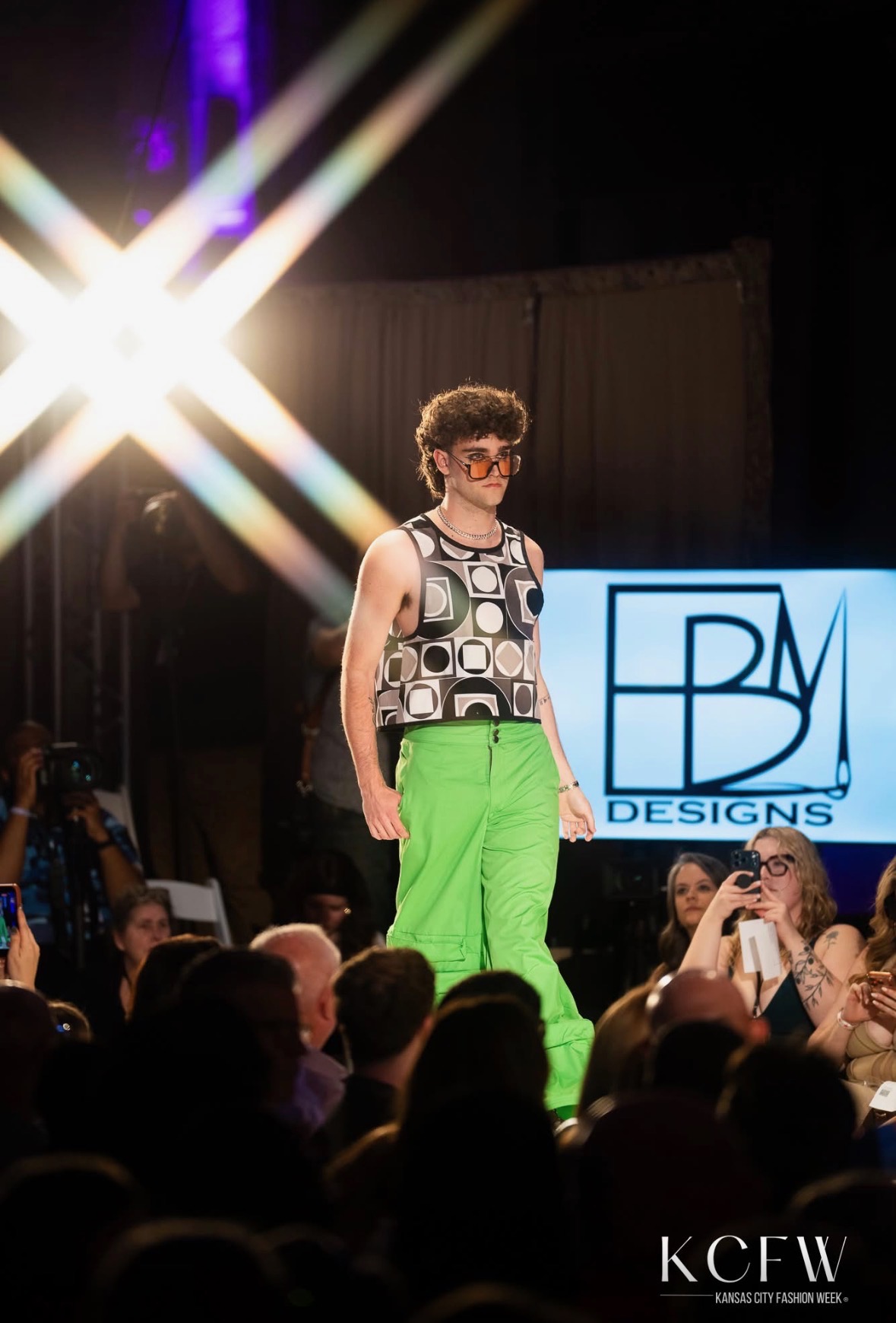 Fashion model walking on runway with audience in foreground, bright stage lights, and a screen displaying 'BDM DESIGNS' in background.