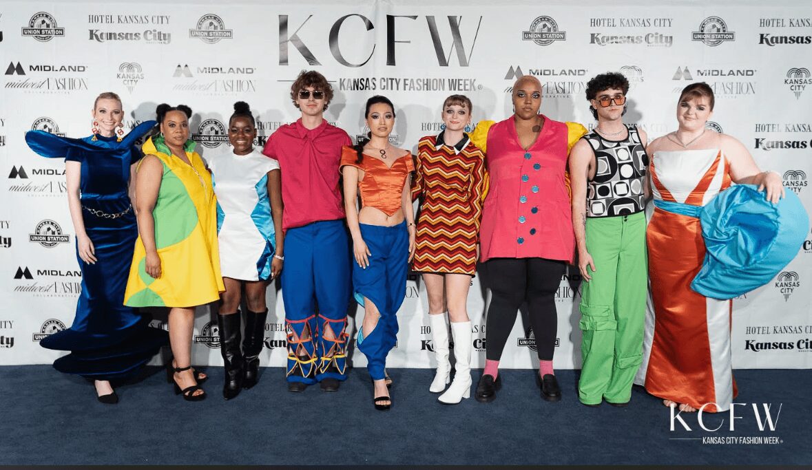 Group of nine people in colorful outfits posing on a blue carpet at Kansas City Fashion Week, backdrop with event logos.