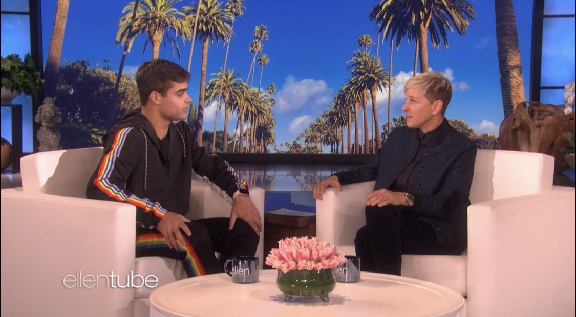 Two people sit on white chairs facing each other with a pink flower arrangement on a table between them, palm trees in background.