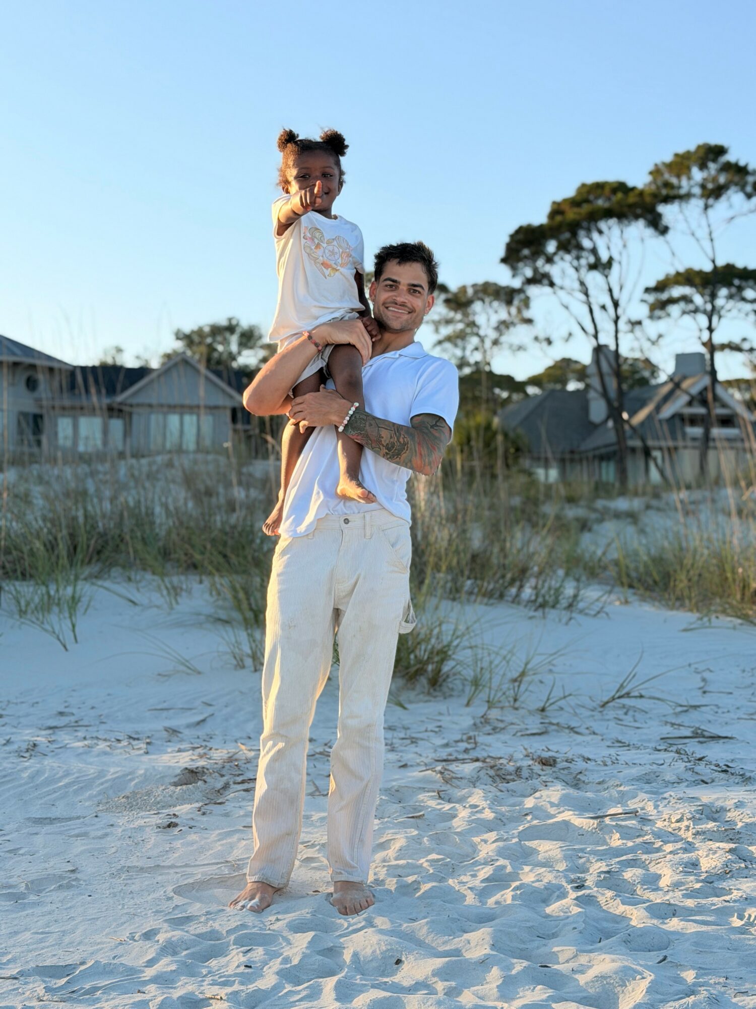 Man carrying a young girl on his shoulders on a sandy beach with houses and trees in the background.