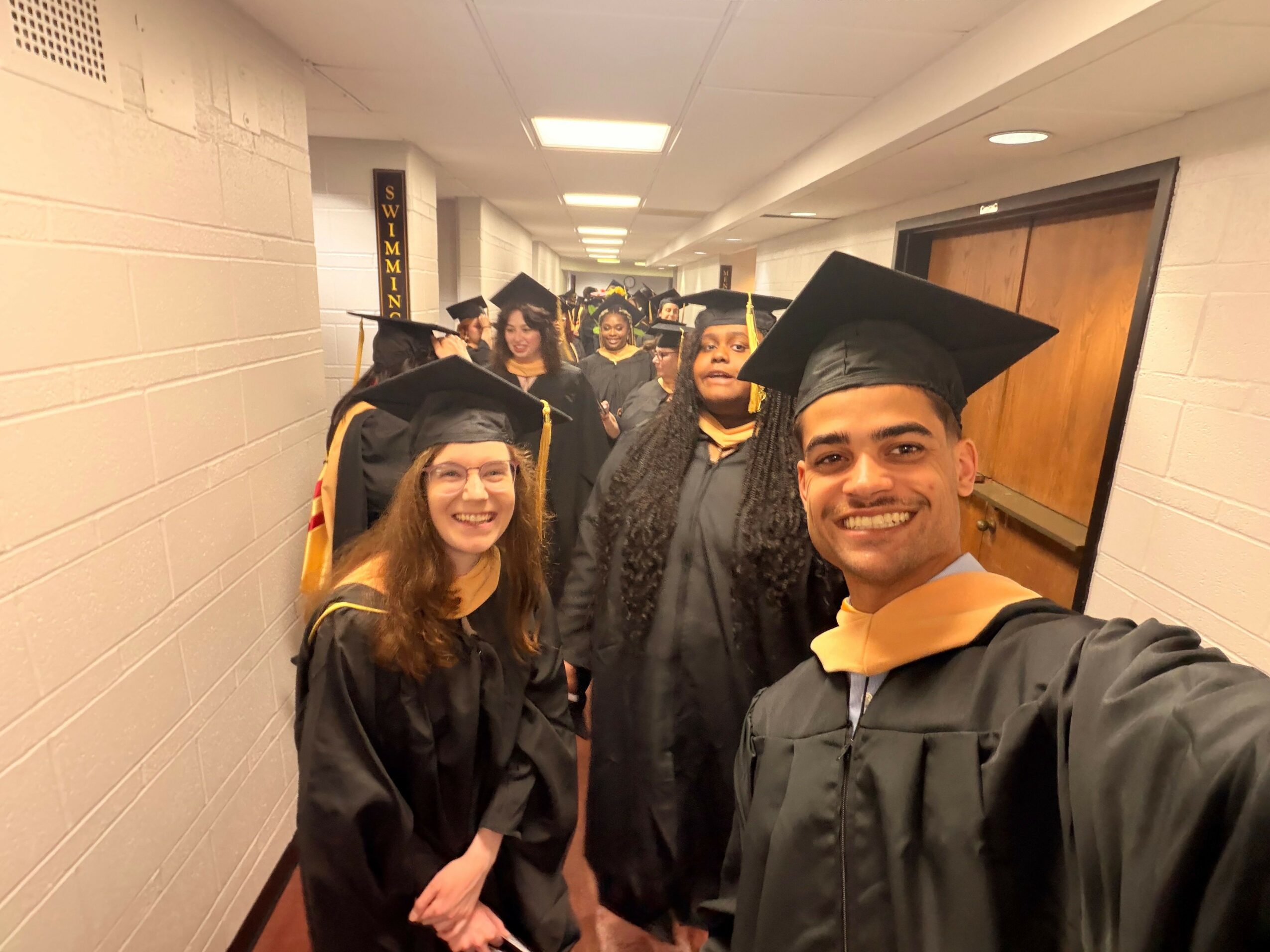 Group of graduates in caps and gowns smiling in hallway, celebrating graduation.