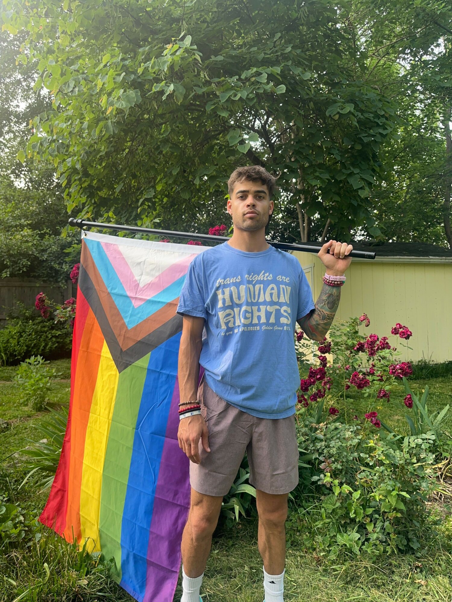 Person holding a rainbow pride flag outdoors with trees and flowers in the background.