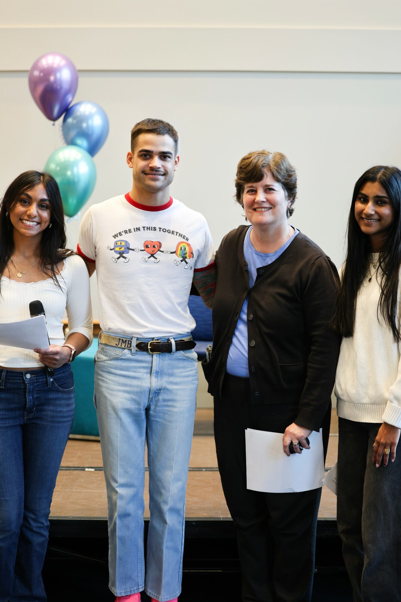 Four people standing together indoors, smiling, with balloons in the background, holding papers, in casual clothing.
