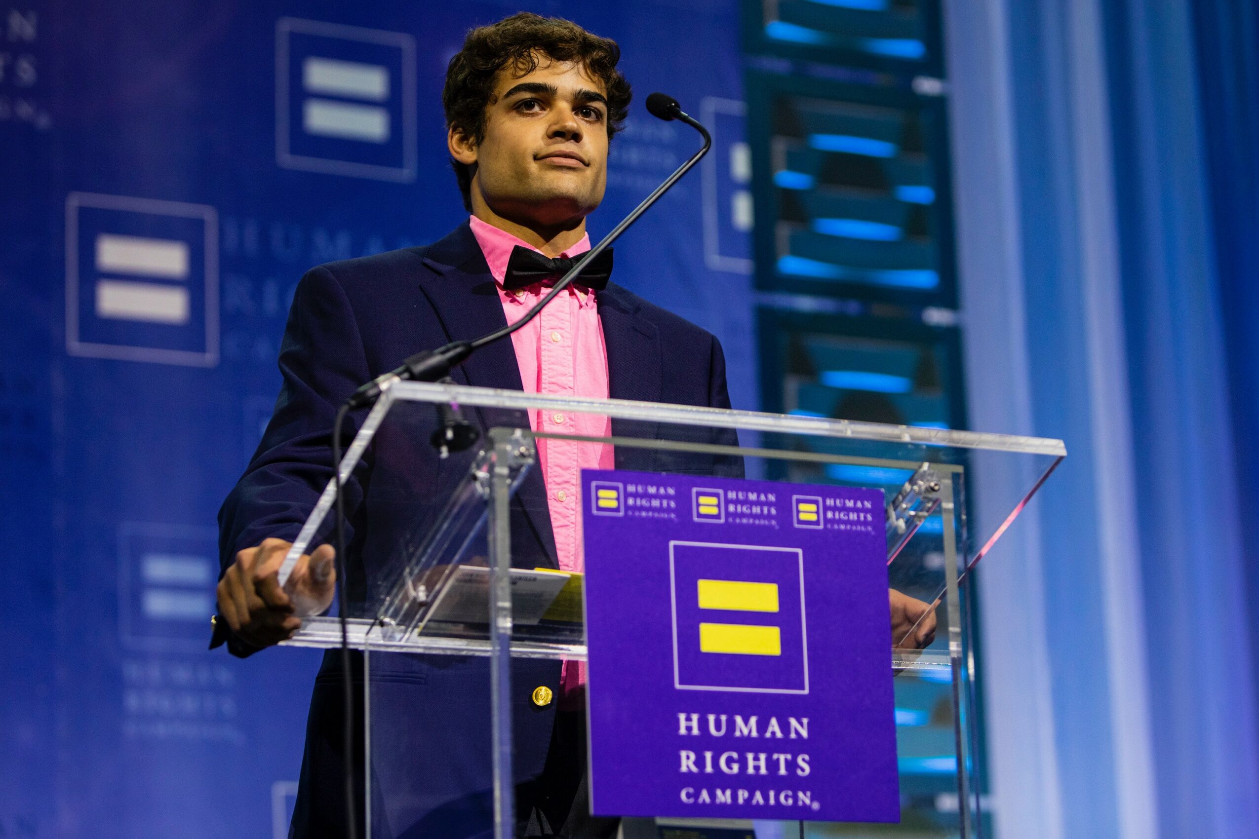 Young man speaking at a podium during a human rights campaign event, with a blue background and logos. Word count: 20