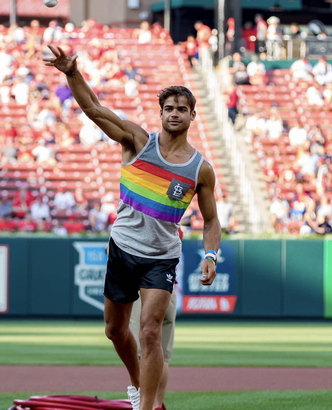 Male athlete in rainbow-striped tank top and black shorts waving at a stadium track, with spectators in the background.