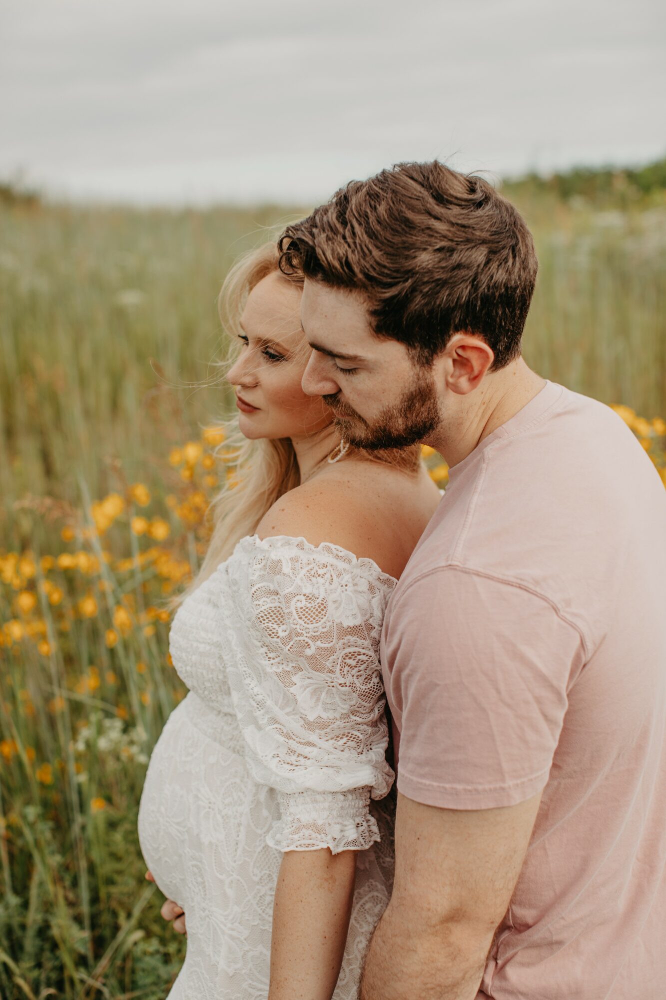 A couple embraces in a field of yellow flowers, with the woman wearing a white lace dress and the man in a pink shirt.