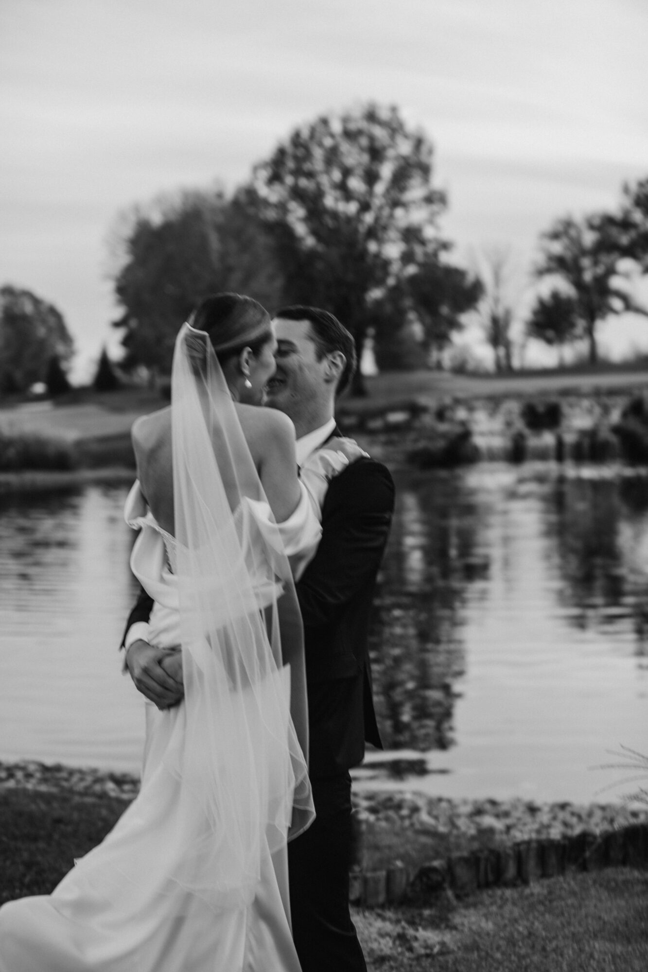 Bride and groom embrace near a river with trees in the background, black and white photo, romantic scene.