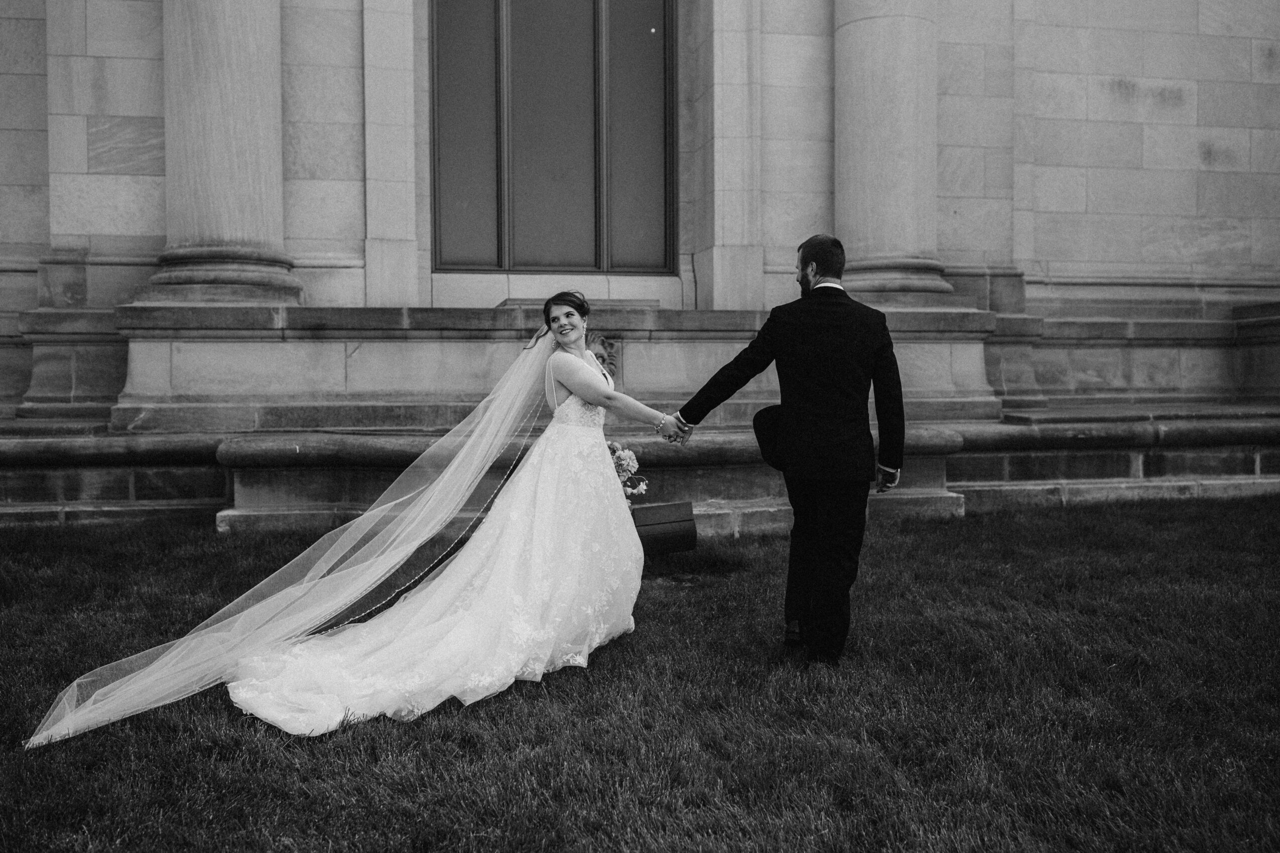 Bride and groom holding hands on grass in front of a building, bride in a long gown with a veil, black and white photo.