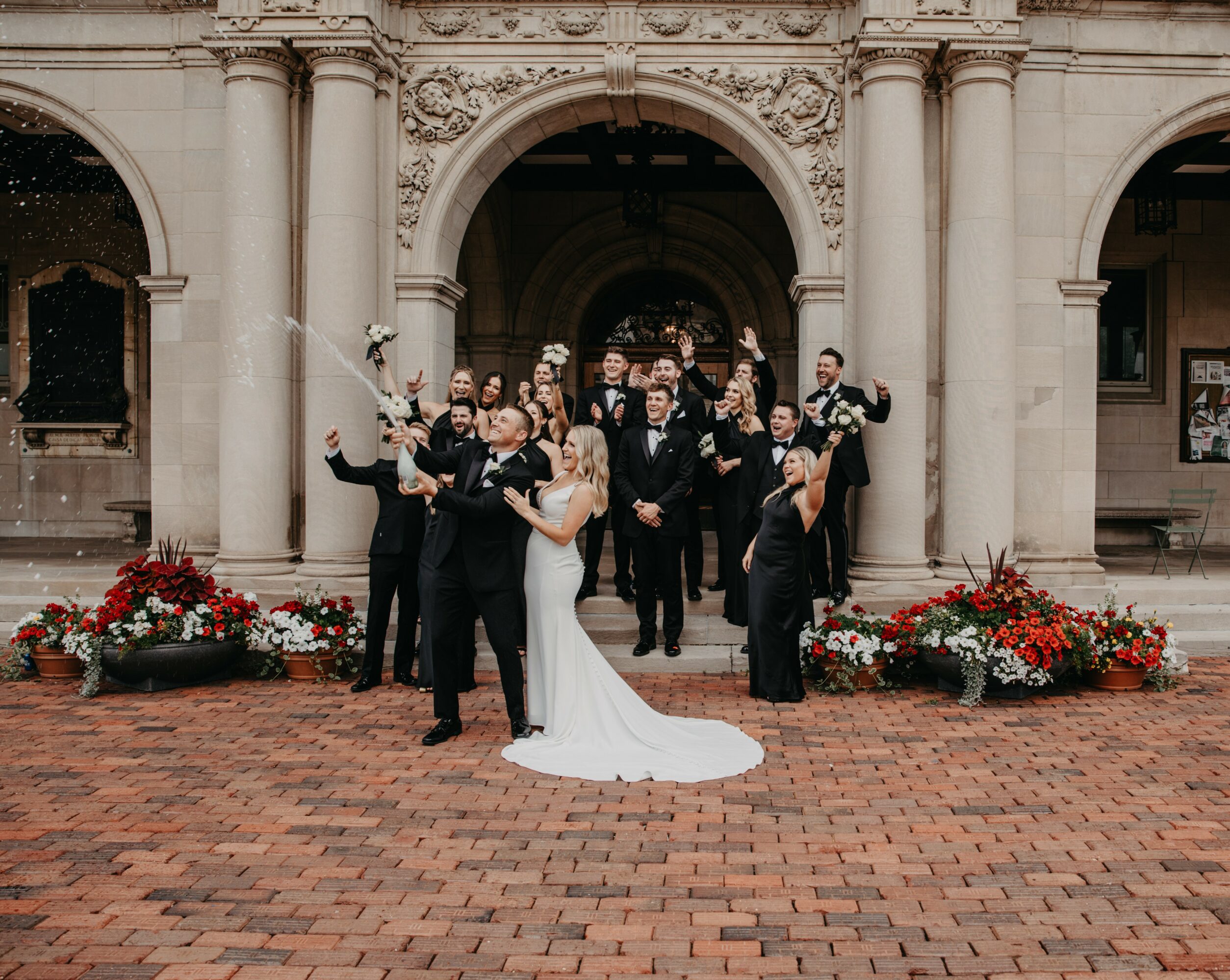 Bride and groom kissing in front of a group of people on a brick courtyard, with floral arrangements and an ornate building entrance.