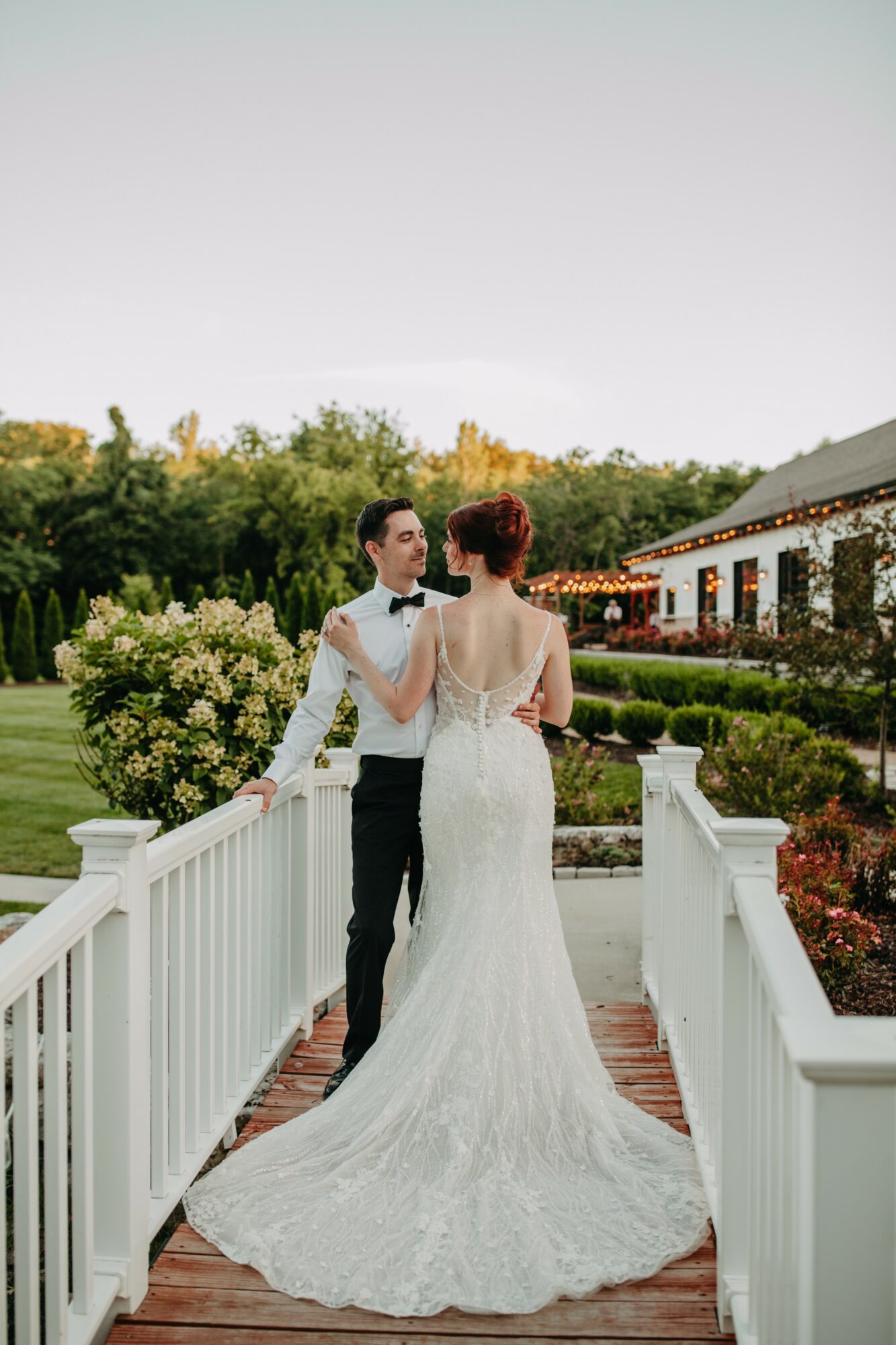 A bride and groom stand on a white bridge outdoors, with trees and a building in the background.