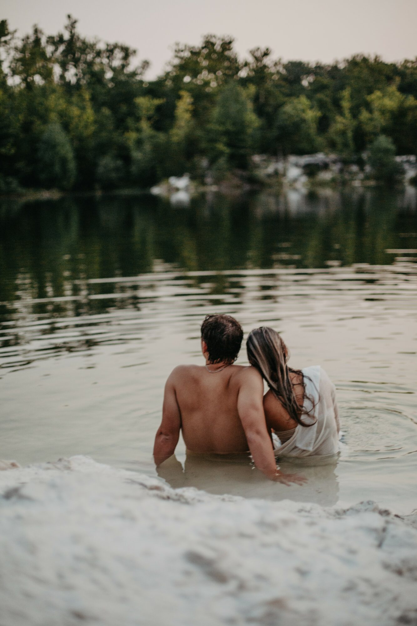 Two people sitting in water near a riverbank with trees in the background, facing away from the camera.