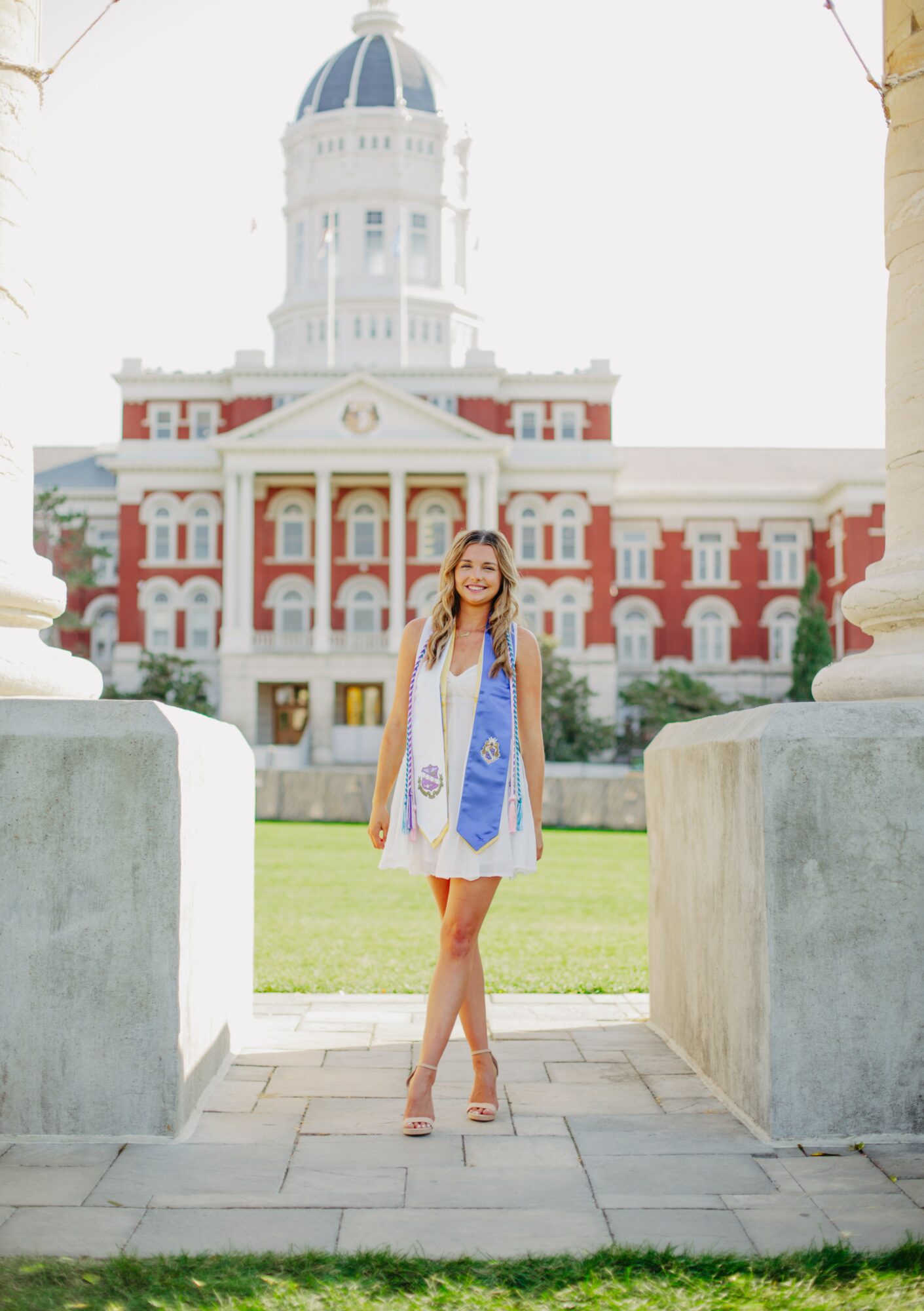 Young woman in a white dress and blue sash standing outdoors in front of a historic building with a dome, columns, and red brick facade.