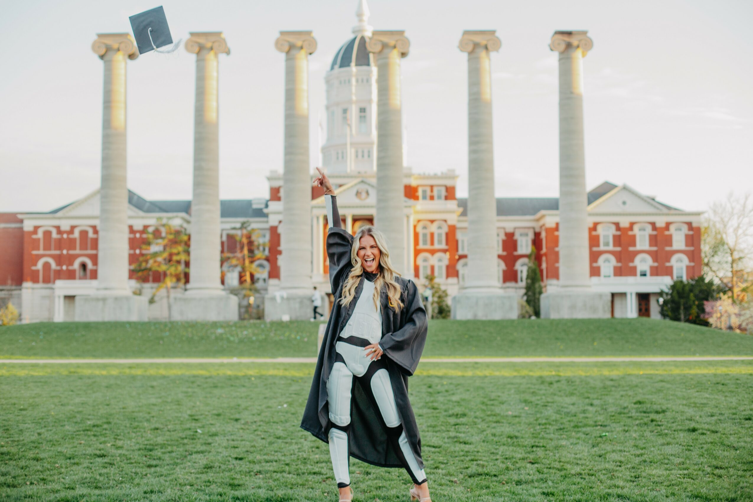 Person in graduation gown and cap standing on grass with historic building and columns in background.