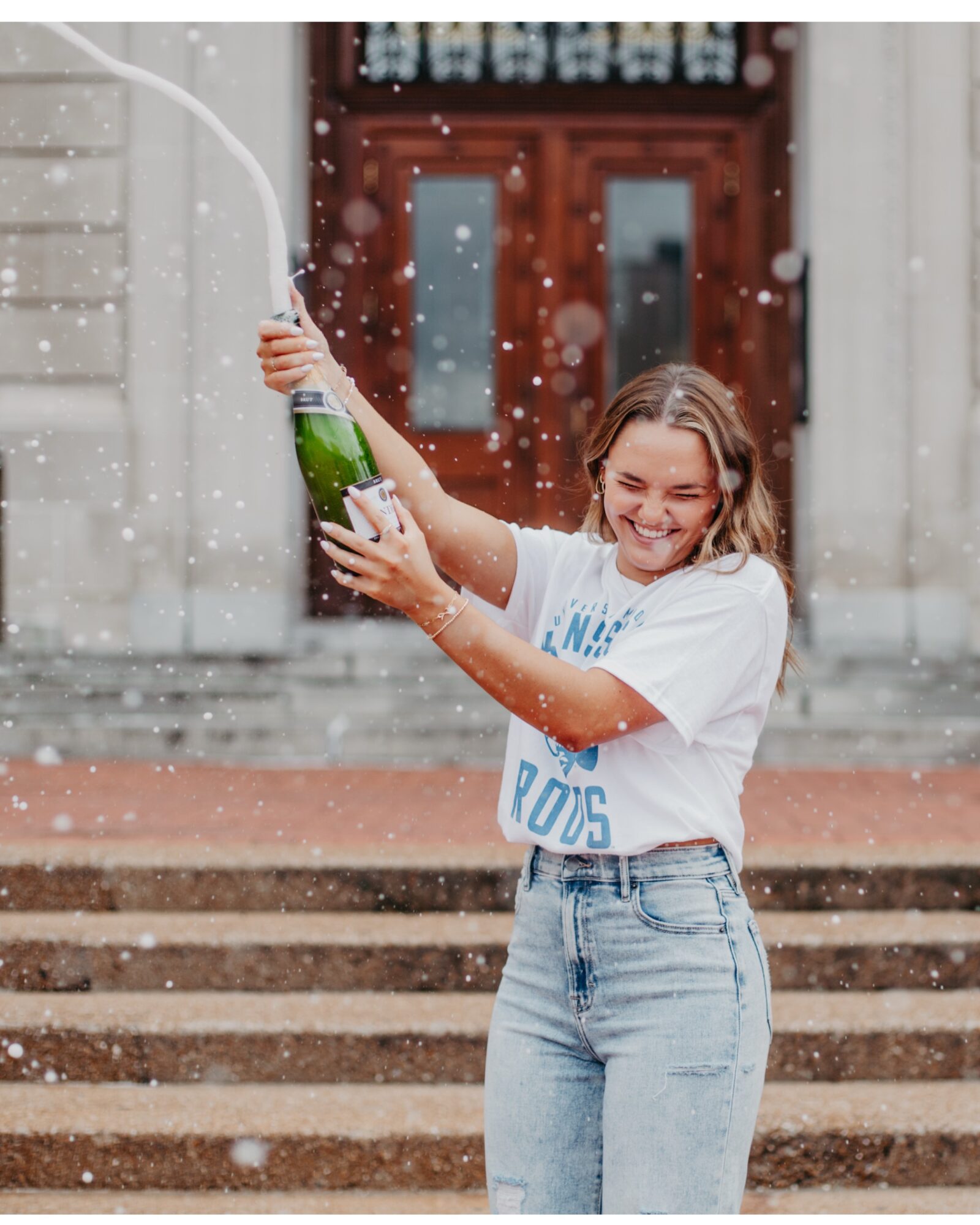 Girl in white T-shirt and jeans holding a champagne bottle, spraying it with water, smiling, in front of a building with steps