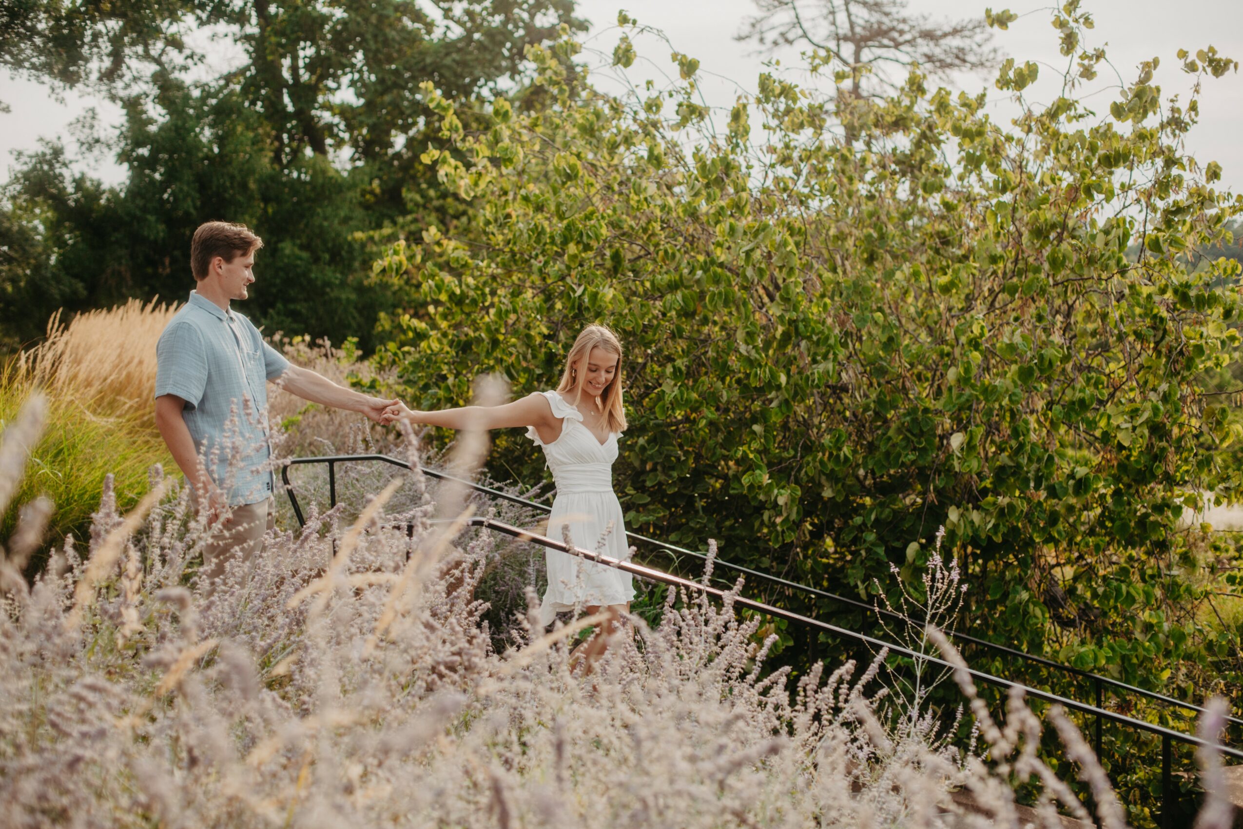 Two people, a man and a woman, hold hands and walk along a path with greenery and flowering plants around them.