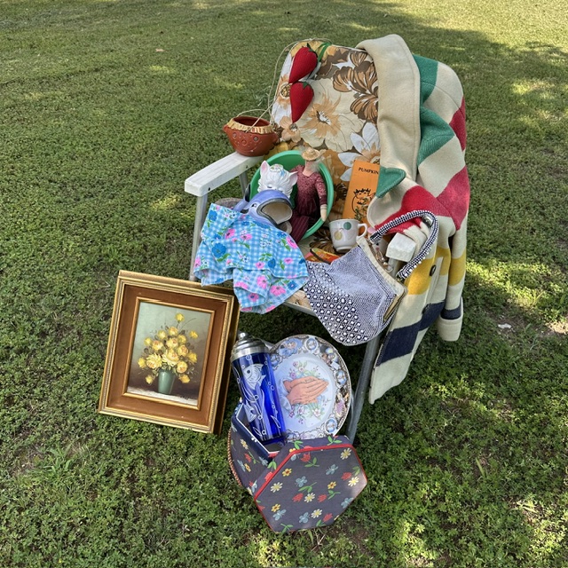 Chair with various items including a framed picture, blanket, bag, and household objects on grass.