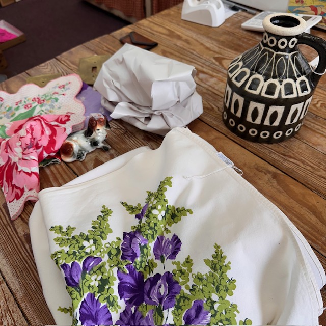 Cloth with purple flowers and green leaves on a wooden table, surrounded by a ceramic jug, crumpled paper, and a small figurine.