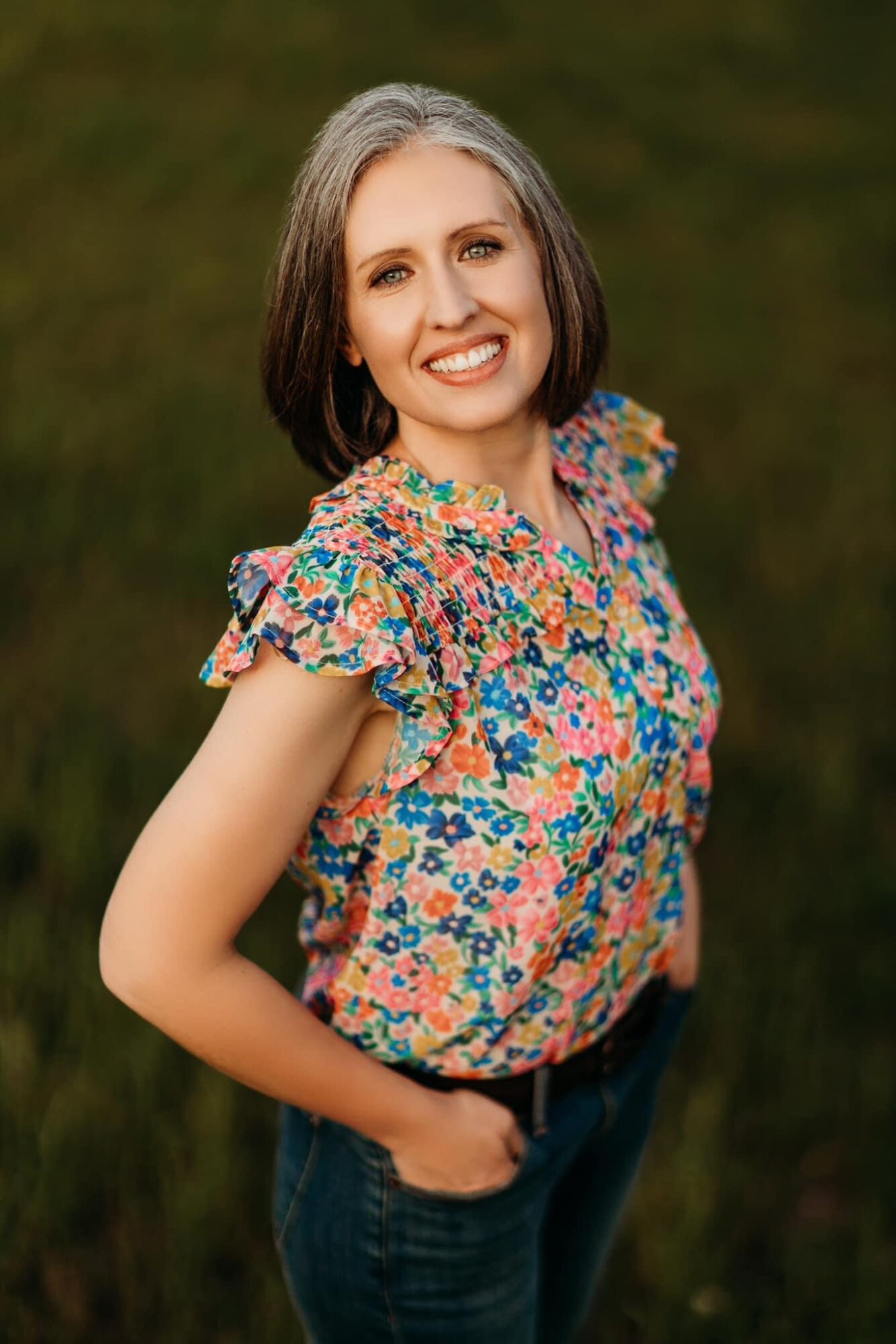 Smiling woman with shoulder-length hair wearing a colorful floral blouse outdoors, hands in pockets, green background.