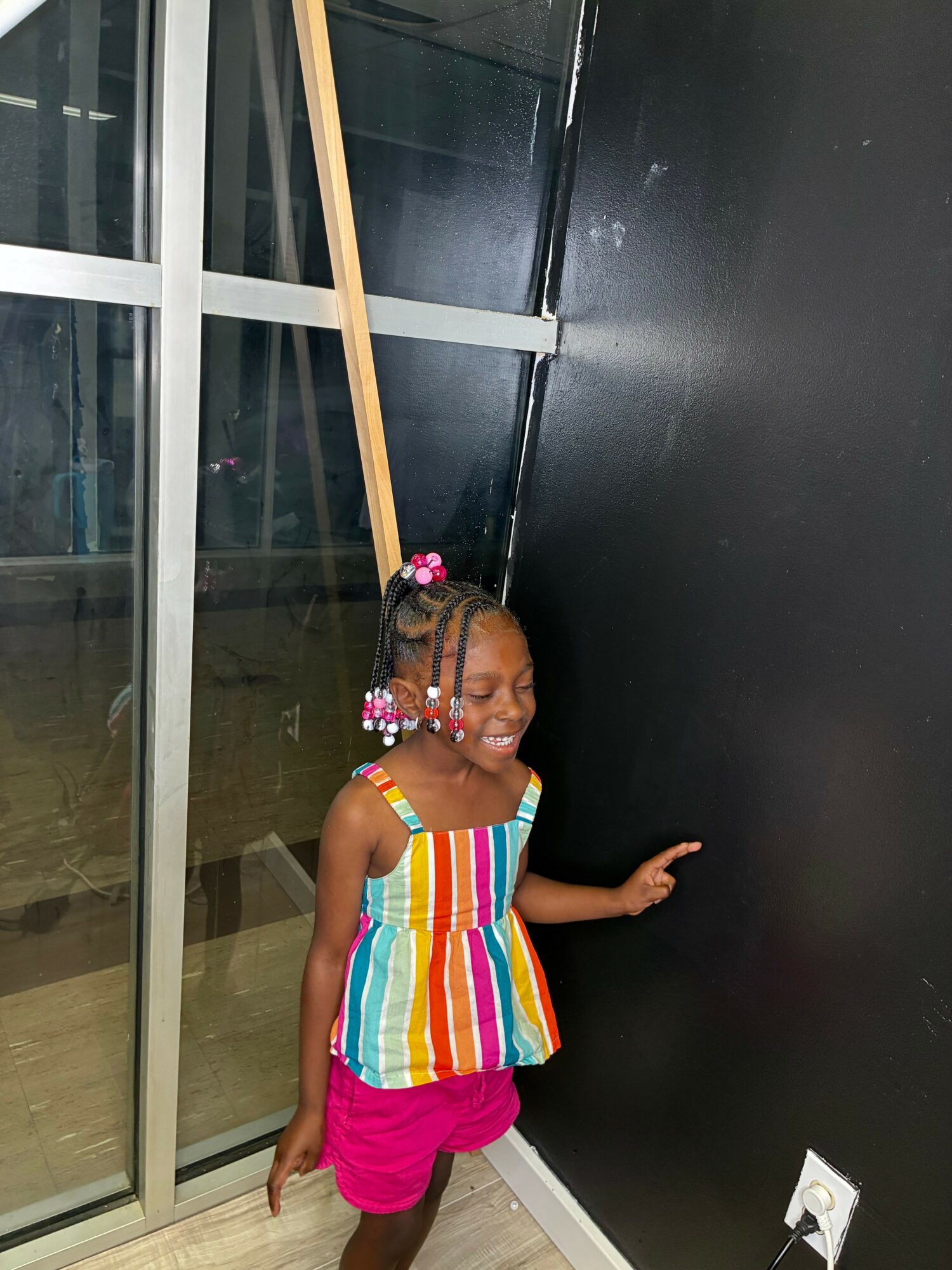 Child with braided hair and colorful beads, wearing a striped dress and pink shorts, standing indoors near a glass door.