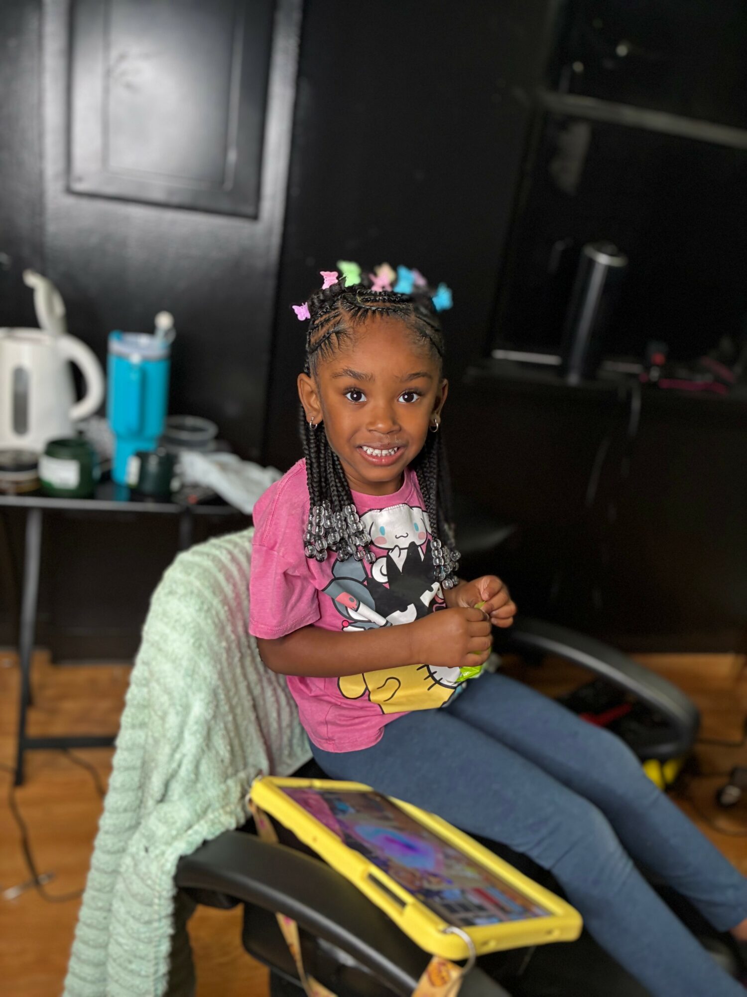 Smiling girl with braided hair and colorful hair accessories, sitting on a chair with a tablet in front, in a room with kitchen items.