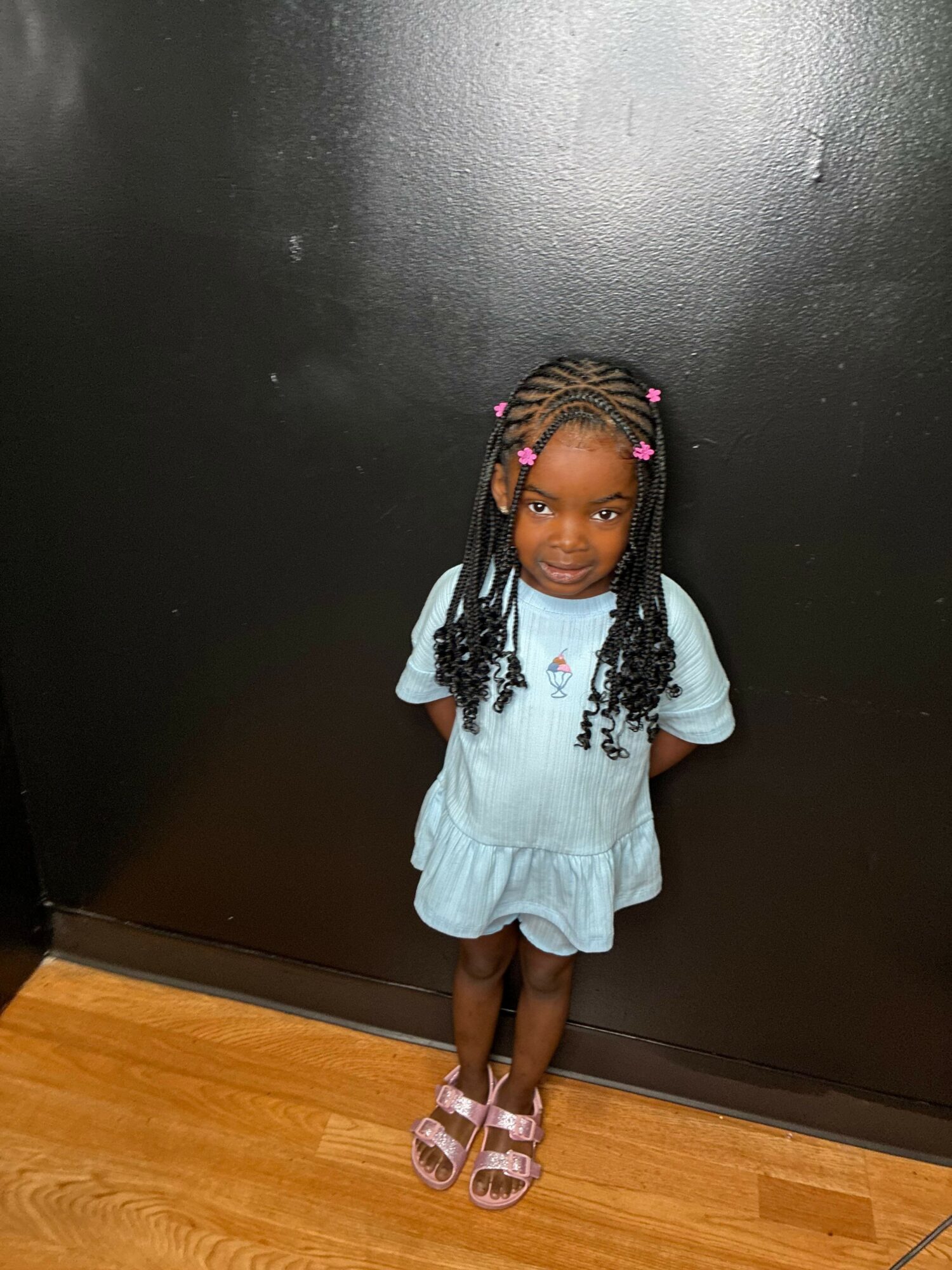 Young girl with braided hair and pink beads, wearing a light blue dress and pink sandals, standing against a black wall.