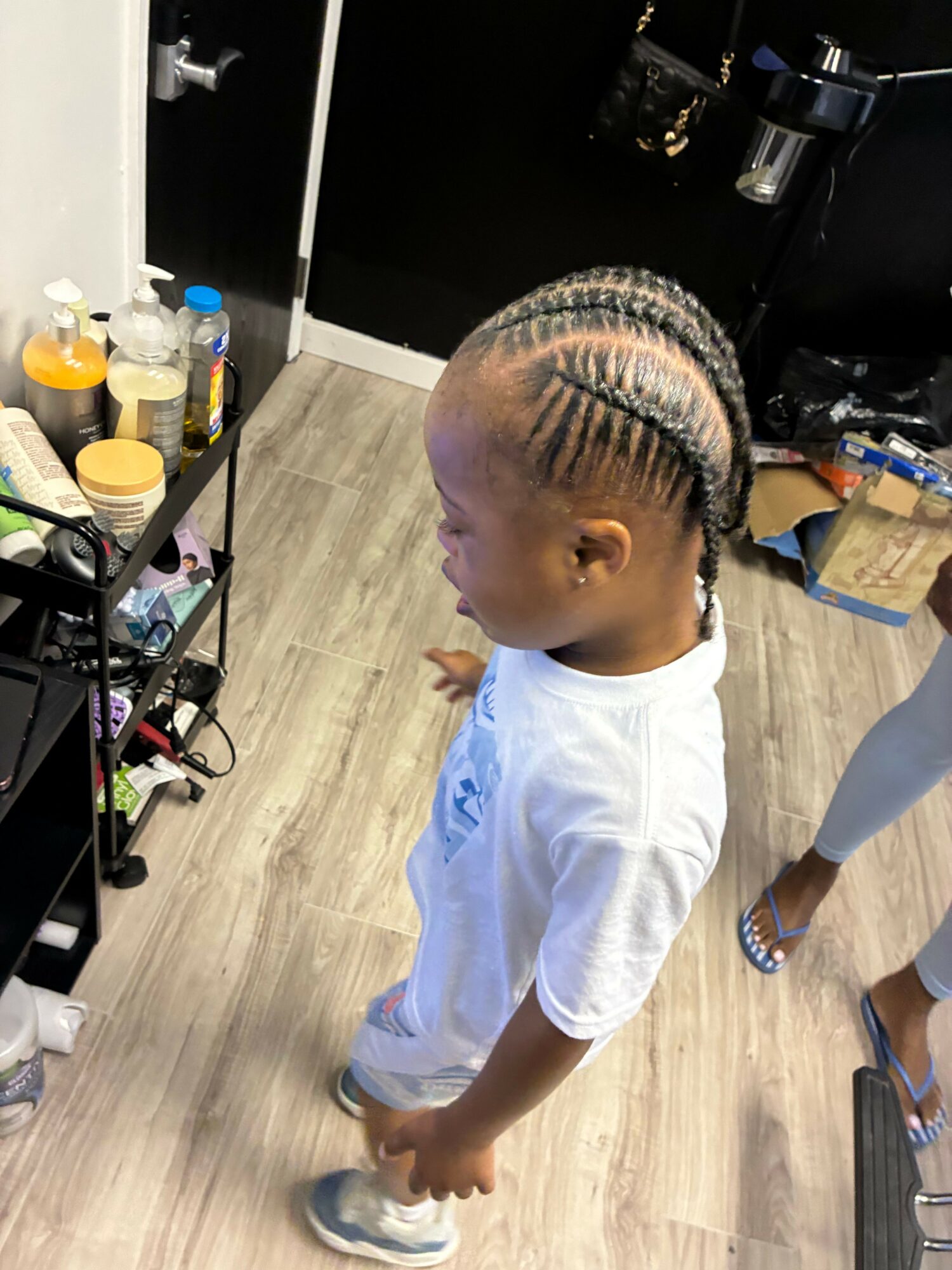 Child with braided hair looking down, standing in a room with wooden floor and various objects nearby.