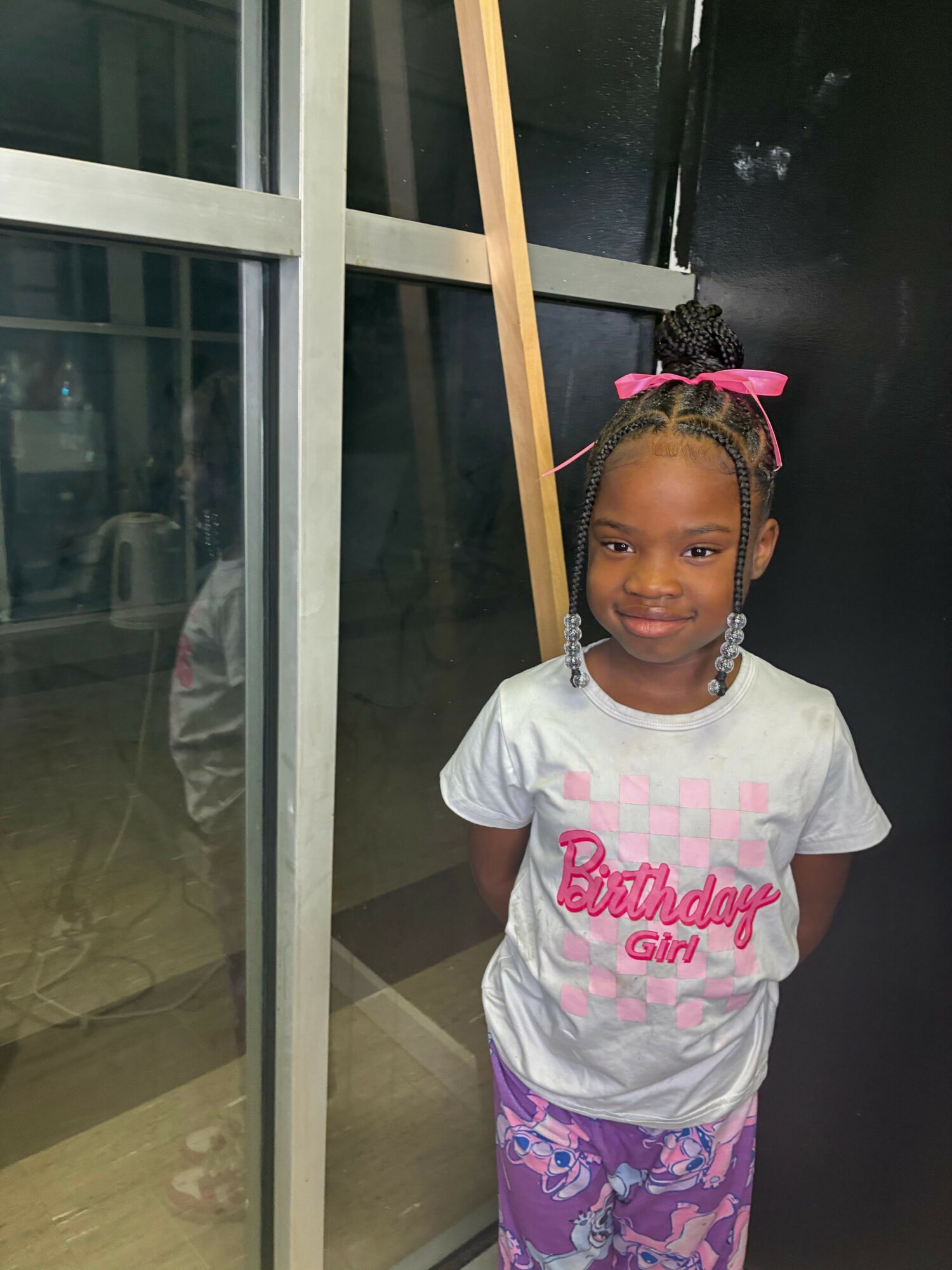 Young girl with braided hair and pink bow, wearing a white t-shirt and colorful pants, standing indoors near a glass window.