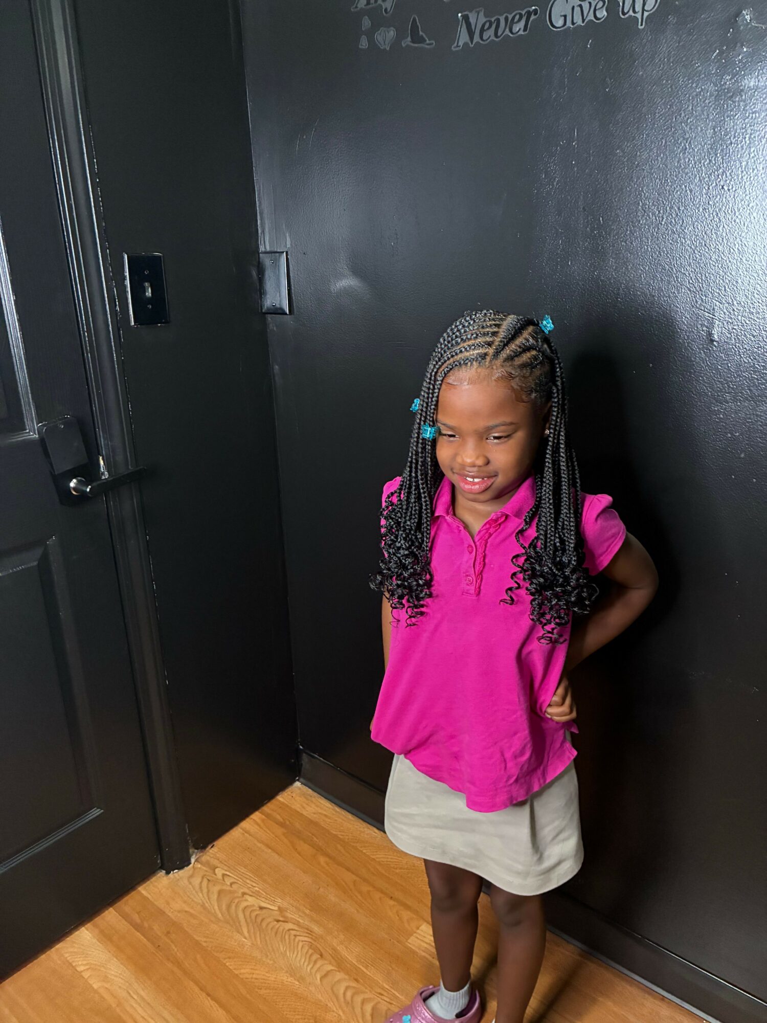 Young girl with braided hair and colorful beads, wearing a pink shirt and gray skirt, standing against a black wall.