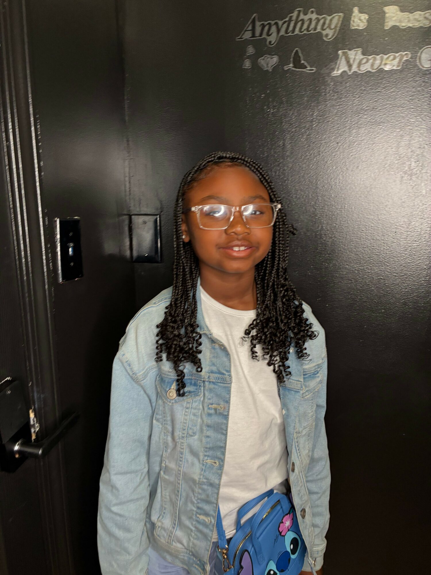Young girl with glasses and braided hair standing against a black wall with writing, wearing a denim jacket and white shirt.