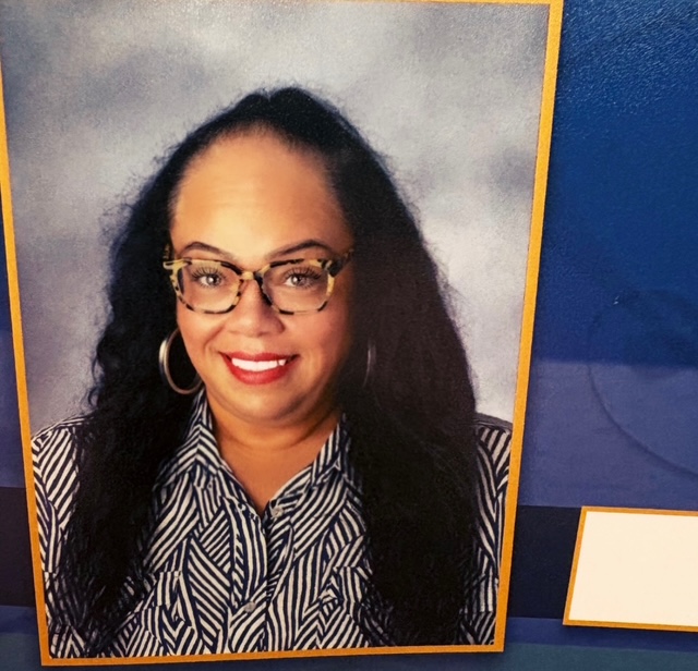 Portrait of a woman with glasses, hoop earrings, and long dark hair, smiling, wearing a patterned shirt.