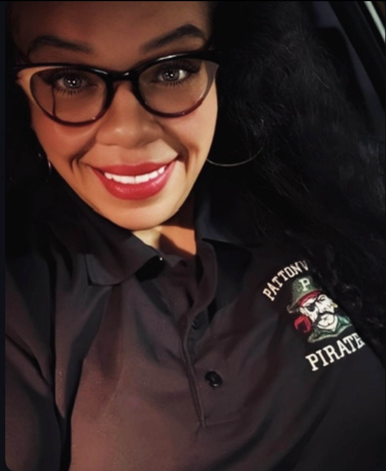 Person with glasses smiling, wearing a black shirt with a pirate logo, in a car.