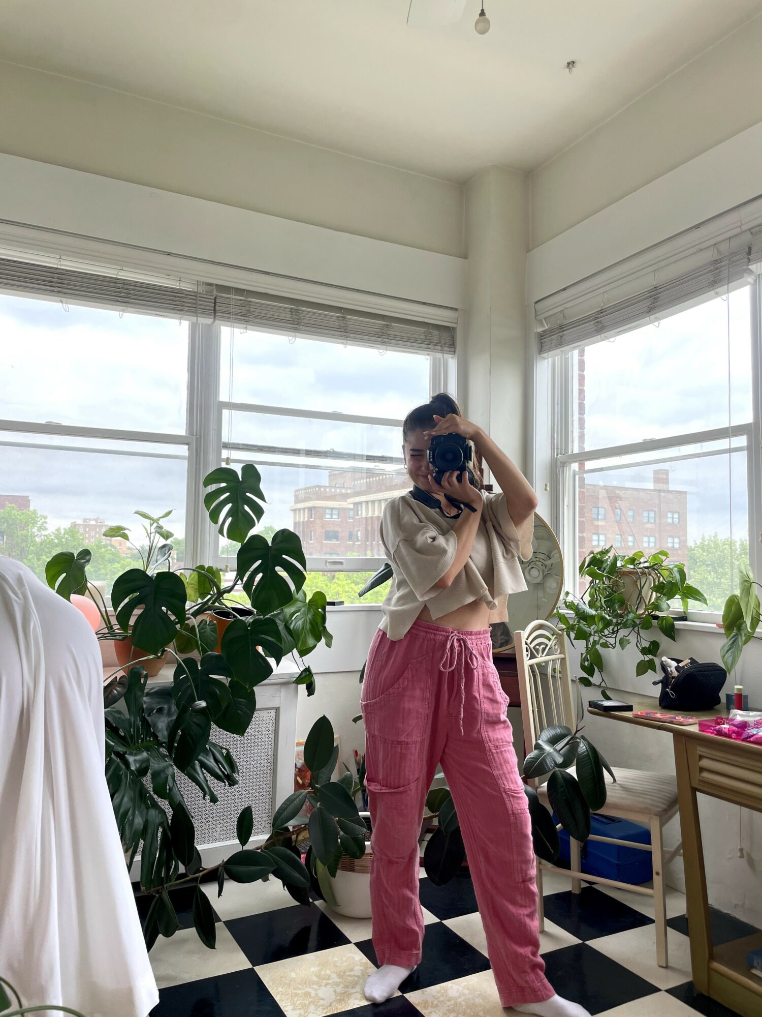 Person taking a mirror selfie in a room with large windows and plants, wearing pink pants and a white top.