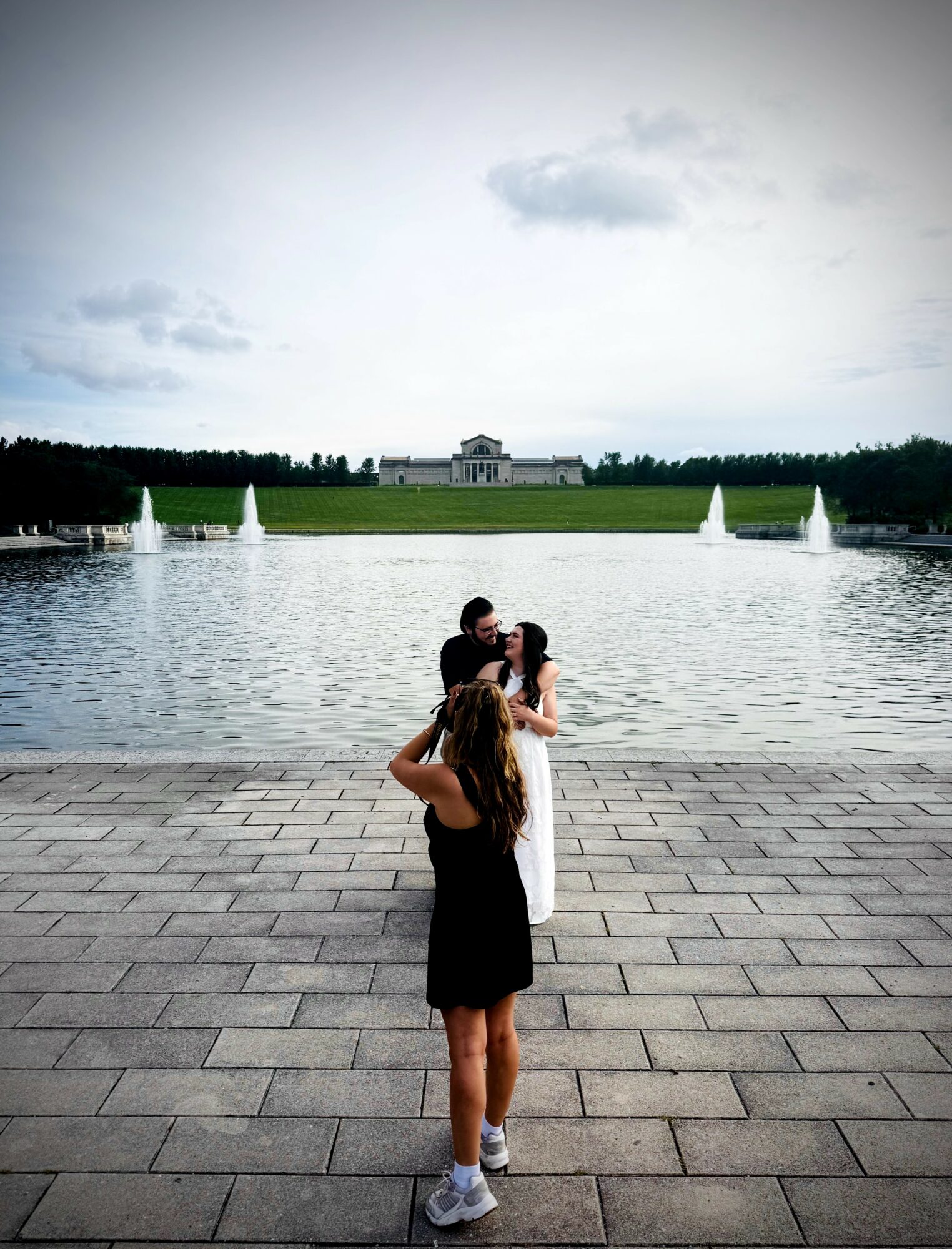 Two women embrace near a large fountain in a park, with a building and cloudy sky in the background.