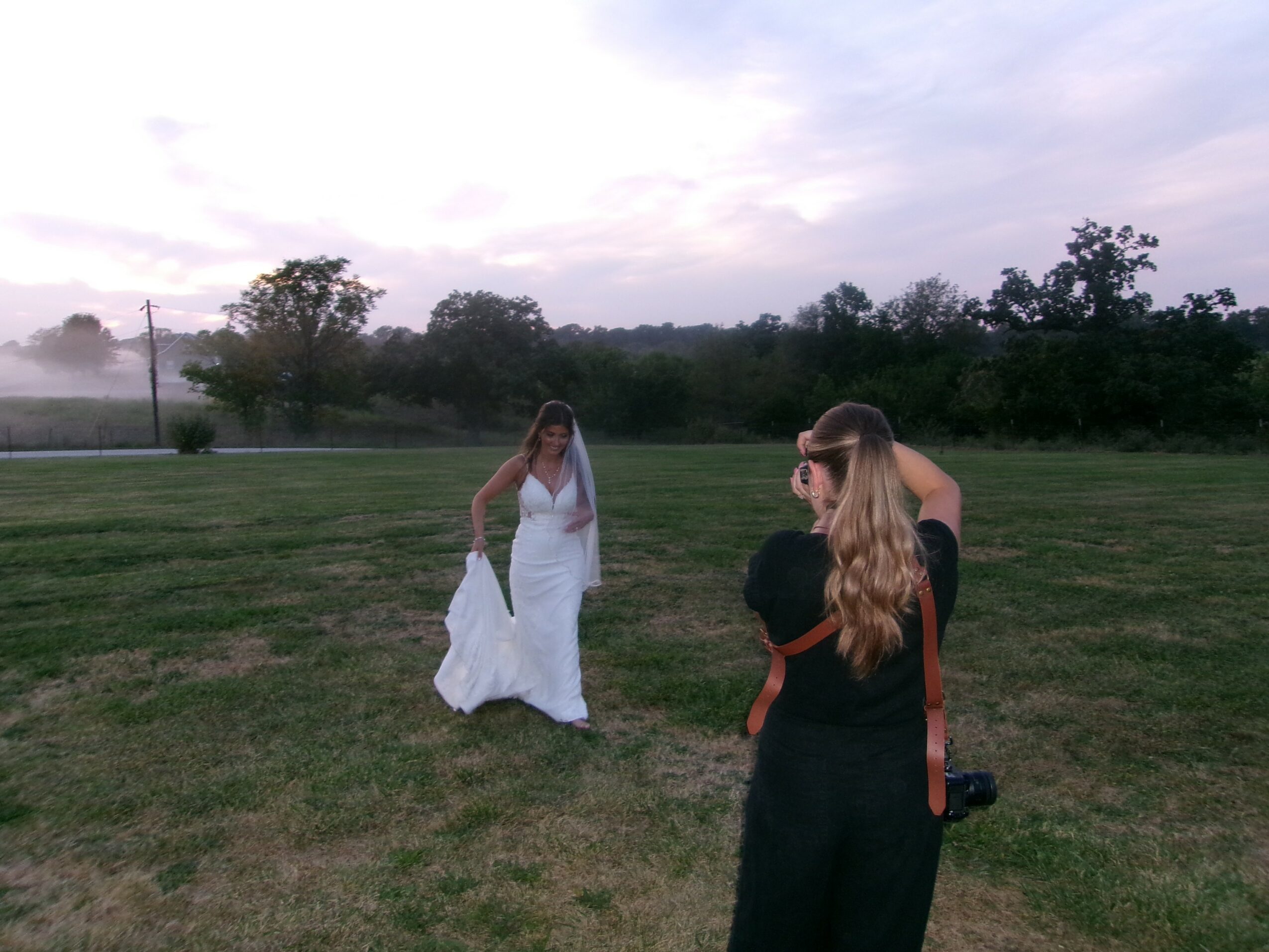 A woman in a white dress stands on grass with trees and sky in the background, a photographer taking her picture.
