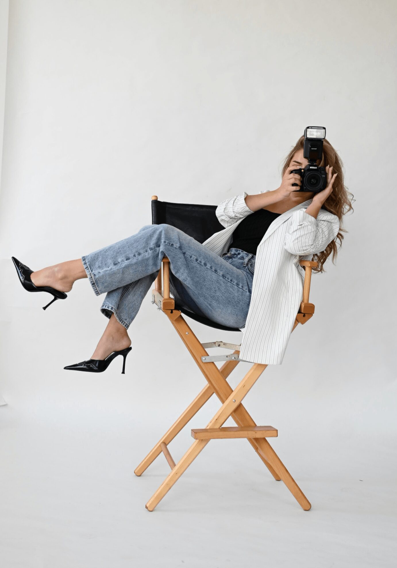 Person sitting on a director's chair, taking a photo with a camera, wearing jeans, a white blazer, and black heels.