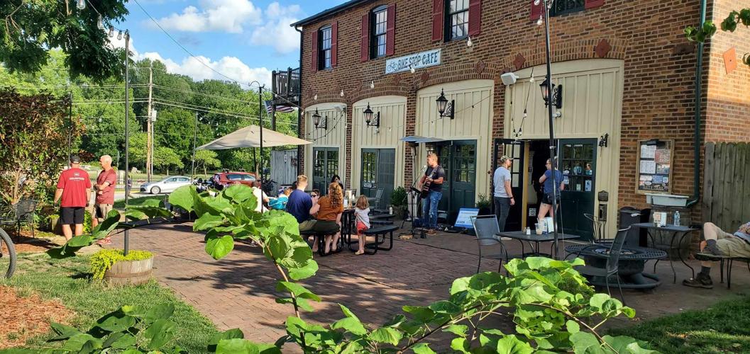 People gather outside a brick building with large windows and outdoor seating, greenery in foreground, sunny day.